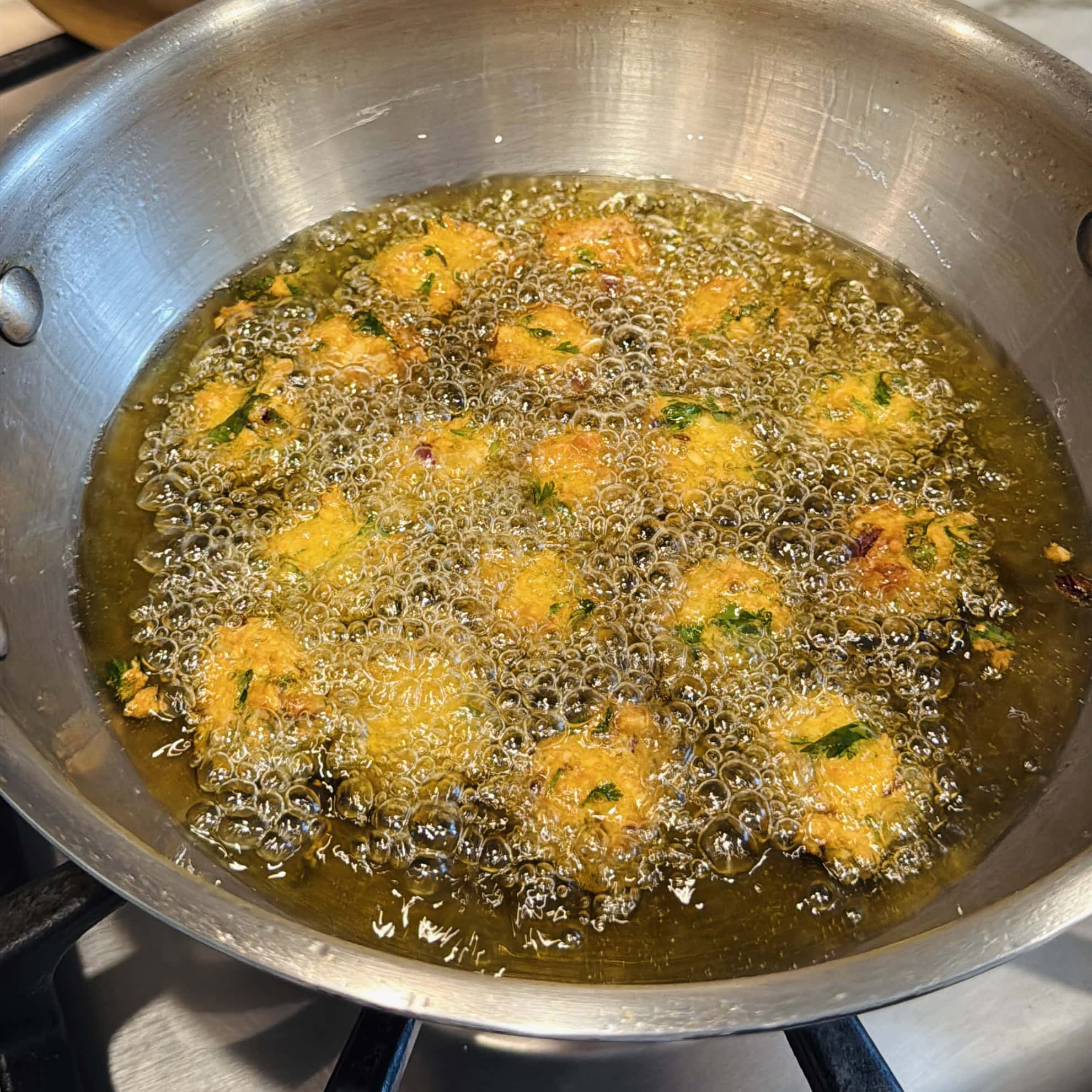Small fritters are being deep-fried in hot oil in a stainless steel pan on a stovetop.