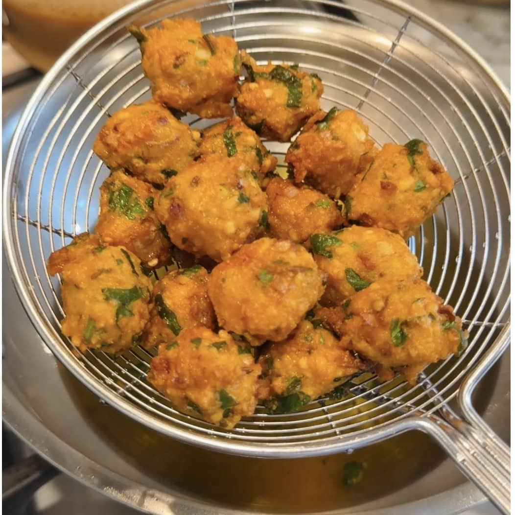 A metal strainer holds several golden-brown, herb-flecked fried balls above a pot of oil.