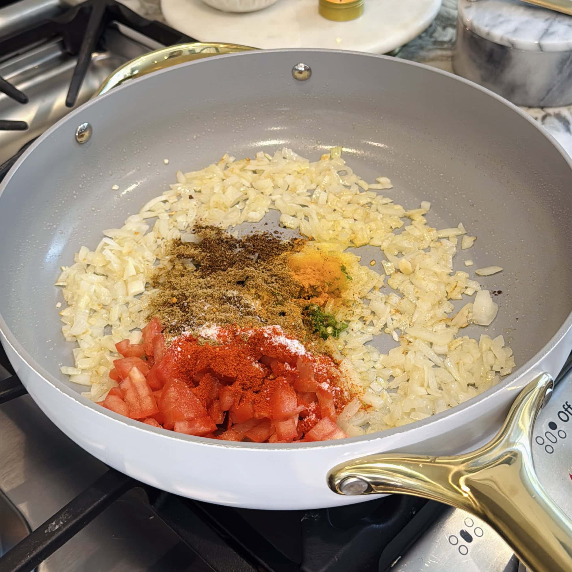 Chopped onions, diced tomatoes, and various ground spices are cooking in a pan on a stovetop.