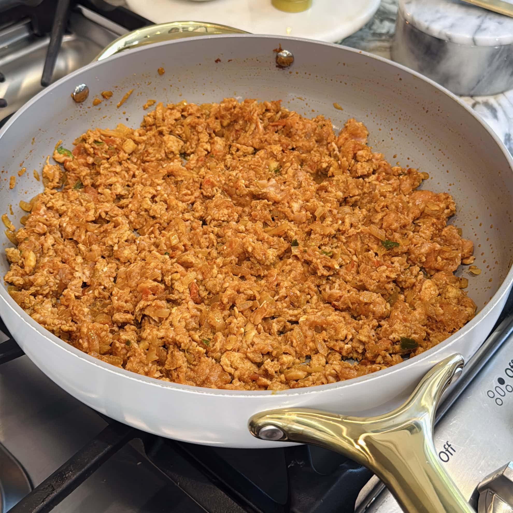 A nonstick skillet on a stove contains cooked, seasoned ground meat mixed with herbs and spices.