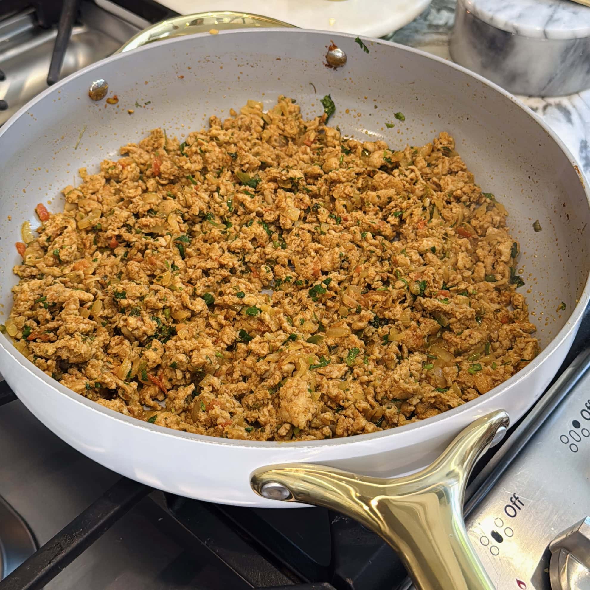 A white skillet with gold handle contains cooked ground kheema mixed with herbs and vegetables on a stovetop.