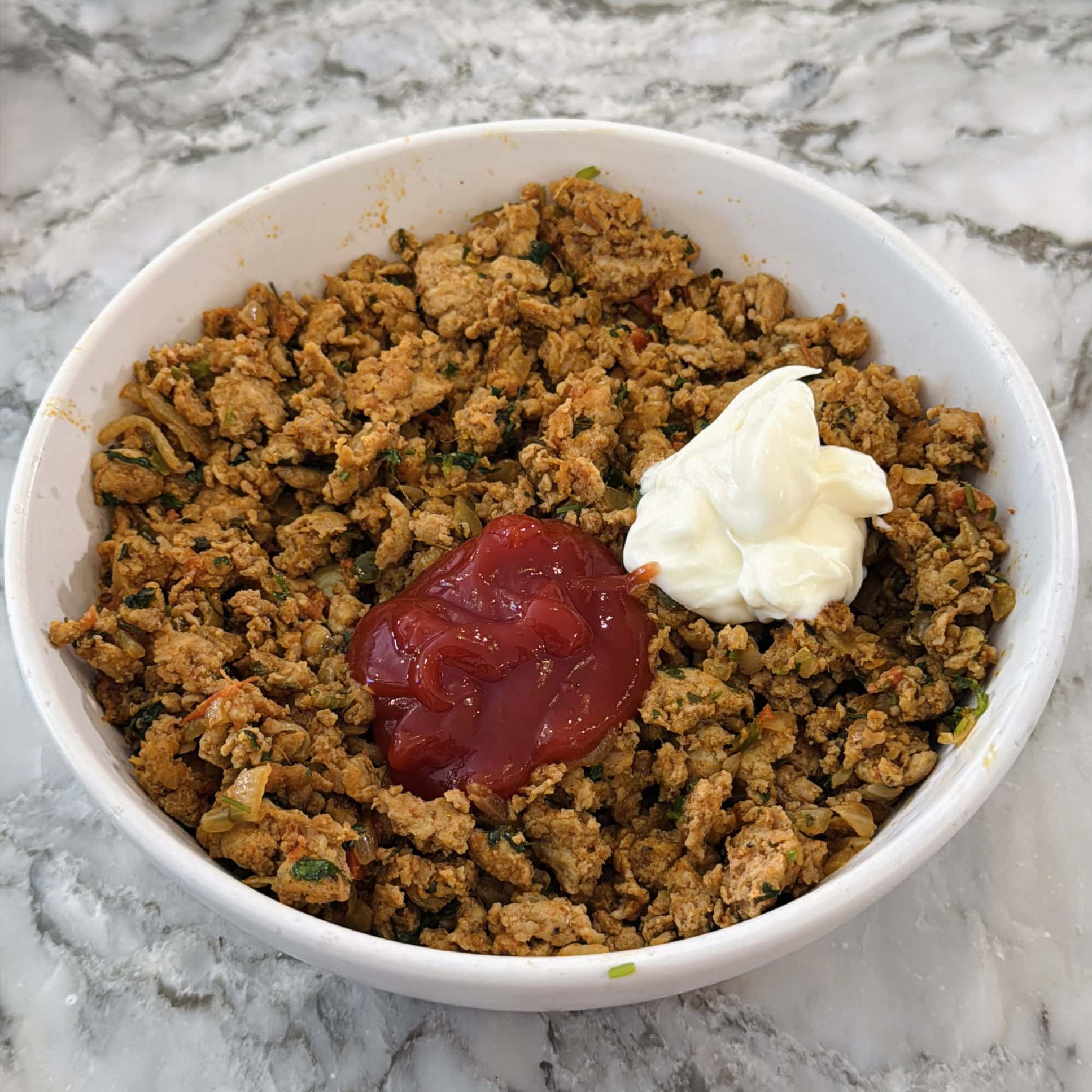A bowl of cooked ground meat with a dollop of ketchup and a dollop of mayonnaise on top, placed on a marble surface.