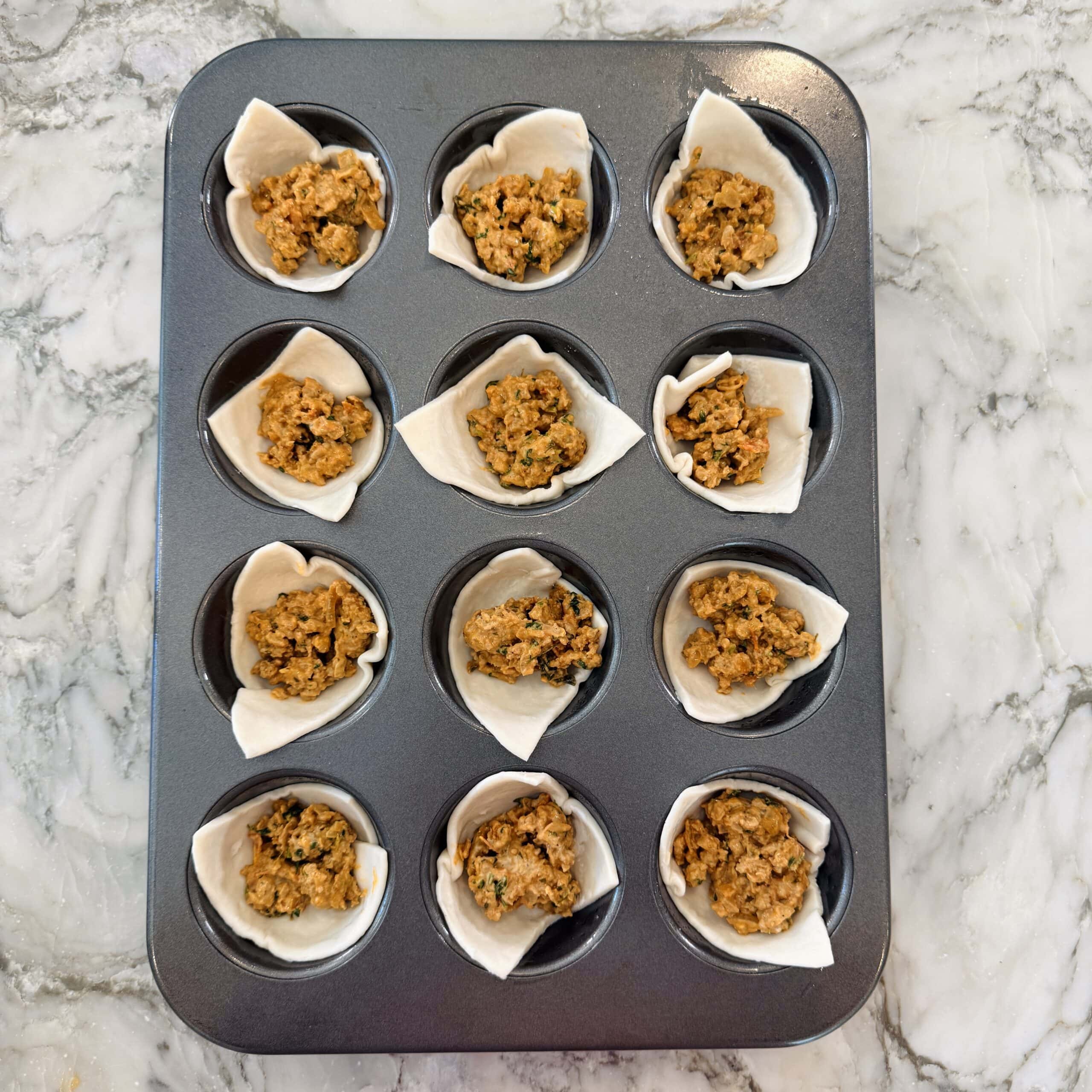 A muffin tin with twelve cups lined with pastry dough squares, each filled with keema.