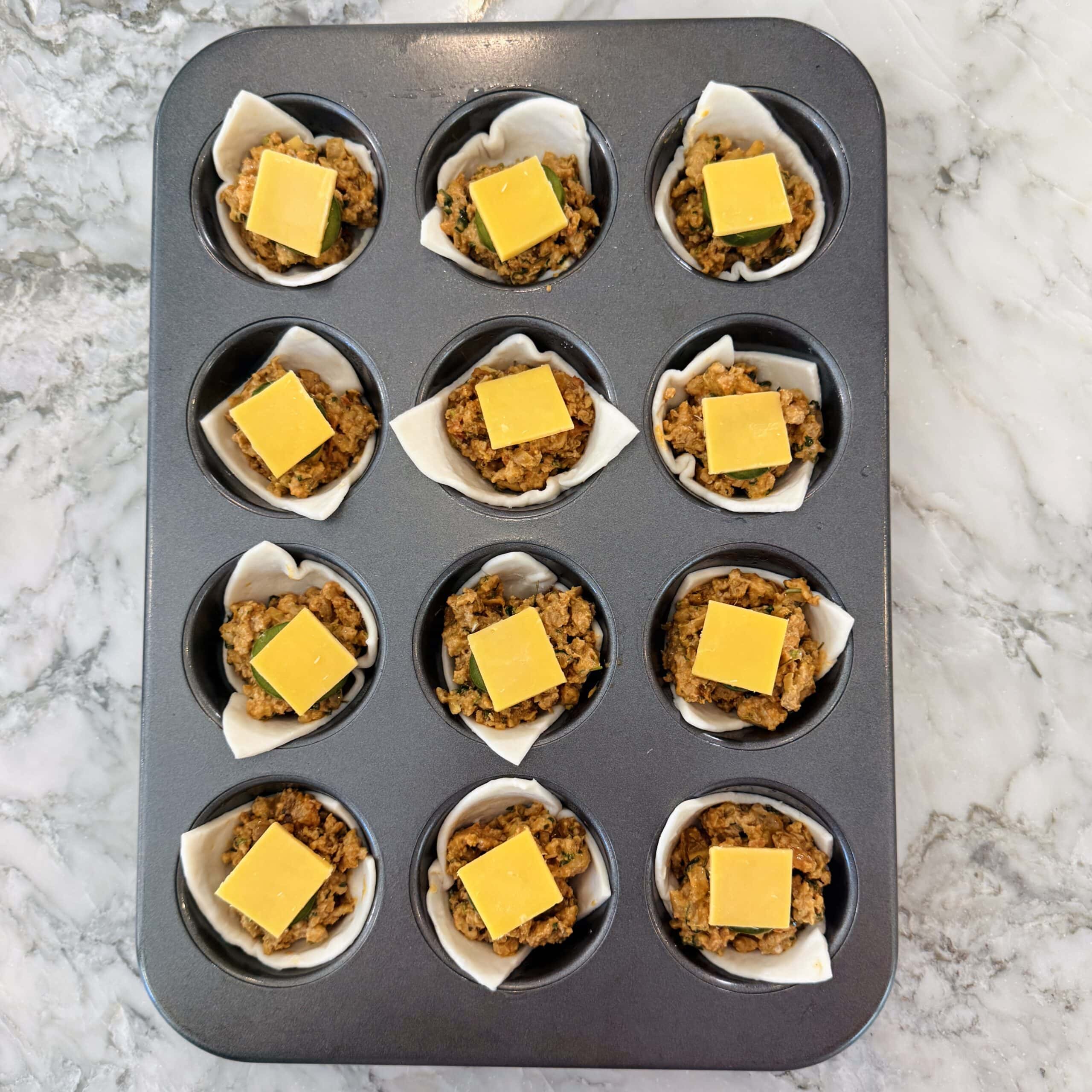 A muffin tin with twelve compartments, each filled with pastry, a savory filling, and topped with a square slice of yellow cheese, on a marble surface.