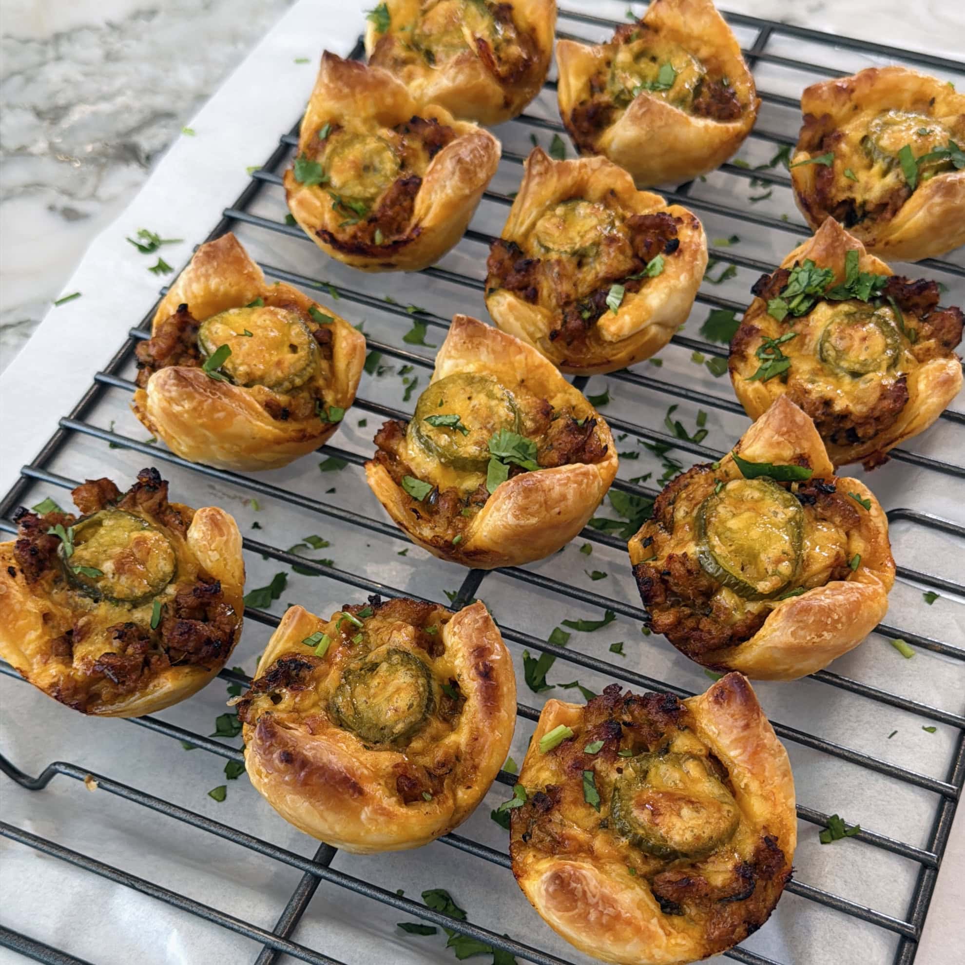Twelve savory puff pastry bites with cheese, herbs, and sliced jalapeños are cooling on a wire rack atop a marble surface.
