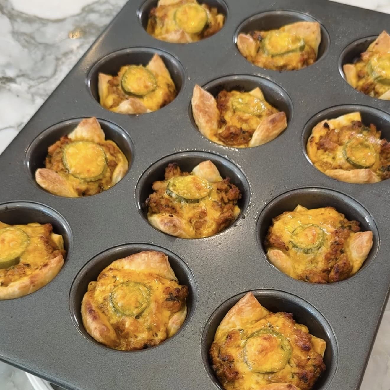 A muffin tin with twelve baked savory pastries, each topped with melted cheese and a slice of jalapeño, sits on a marble countertop.
