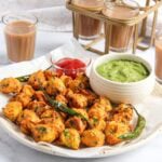 A plate of vegetable pakoras with green chili peppers, served with green chutney and ketchup, alongside glasses of chai tea.