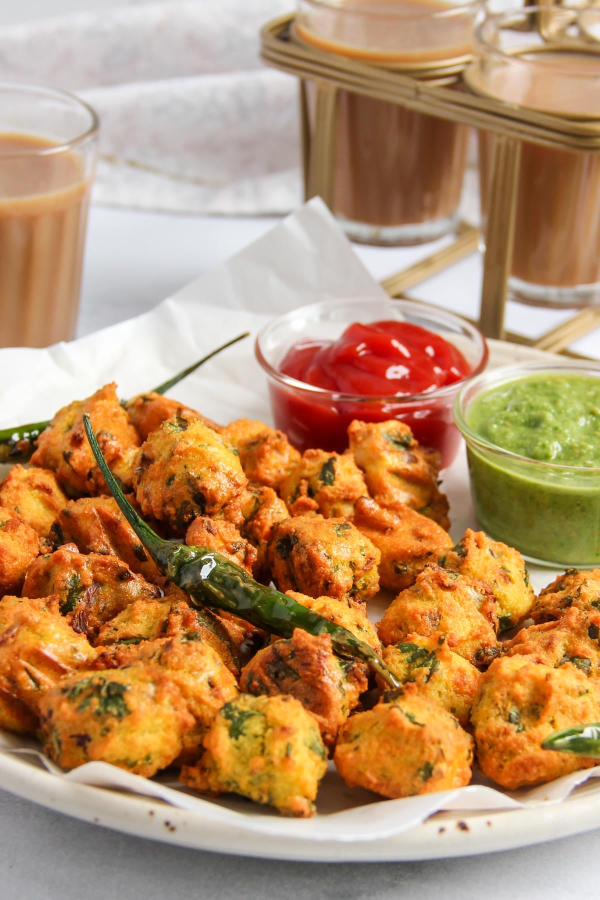 A plate of paneer pakoras garnished with green chilies, served with green chutney and ketchup, with glasses of chai in the background.