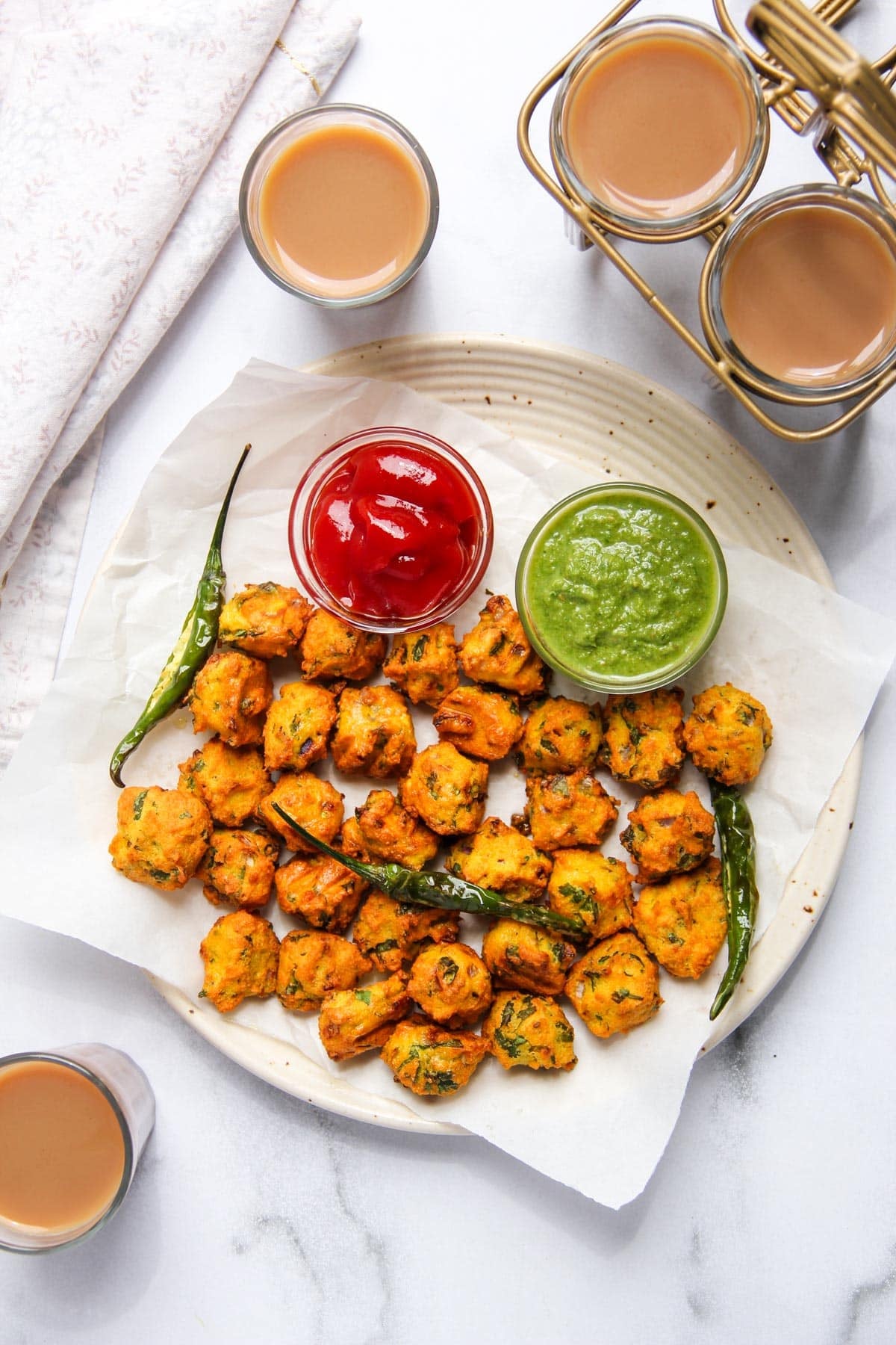 A plate of mung dal bhajiyas with fried green chilies, served with green chutney and ketchup, surrounded by glasses of tea on a white surface.