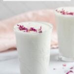 Two glasses of sweet lassi, a yogurt drink, topped with dried rose petals, are placed on a white surface with a pink cloth in the background.