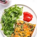 A plate with a slice of baked veggie casserole, fresh arugula salad, and a serving of ketchup, with chia seed pudding in the background.