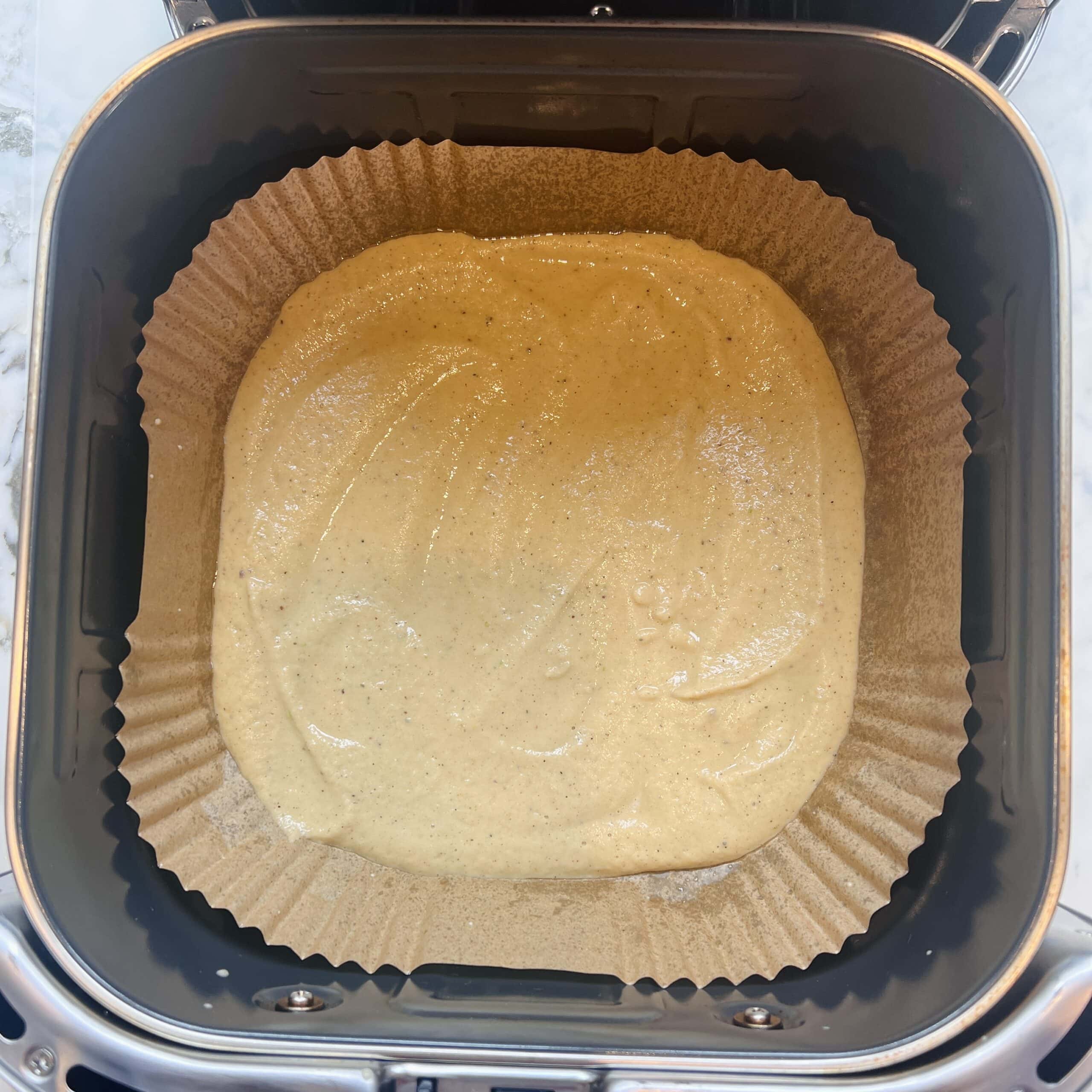 A baking pan lined with parchment paper holds a layer of light brown batter inside an air fryer basket.