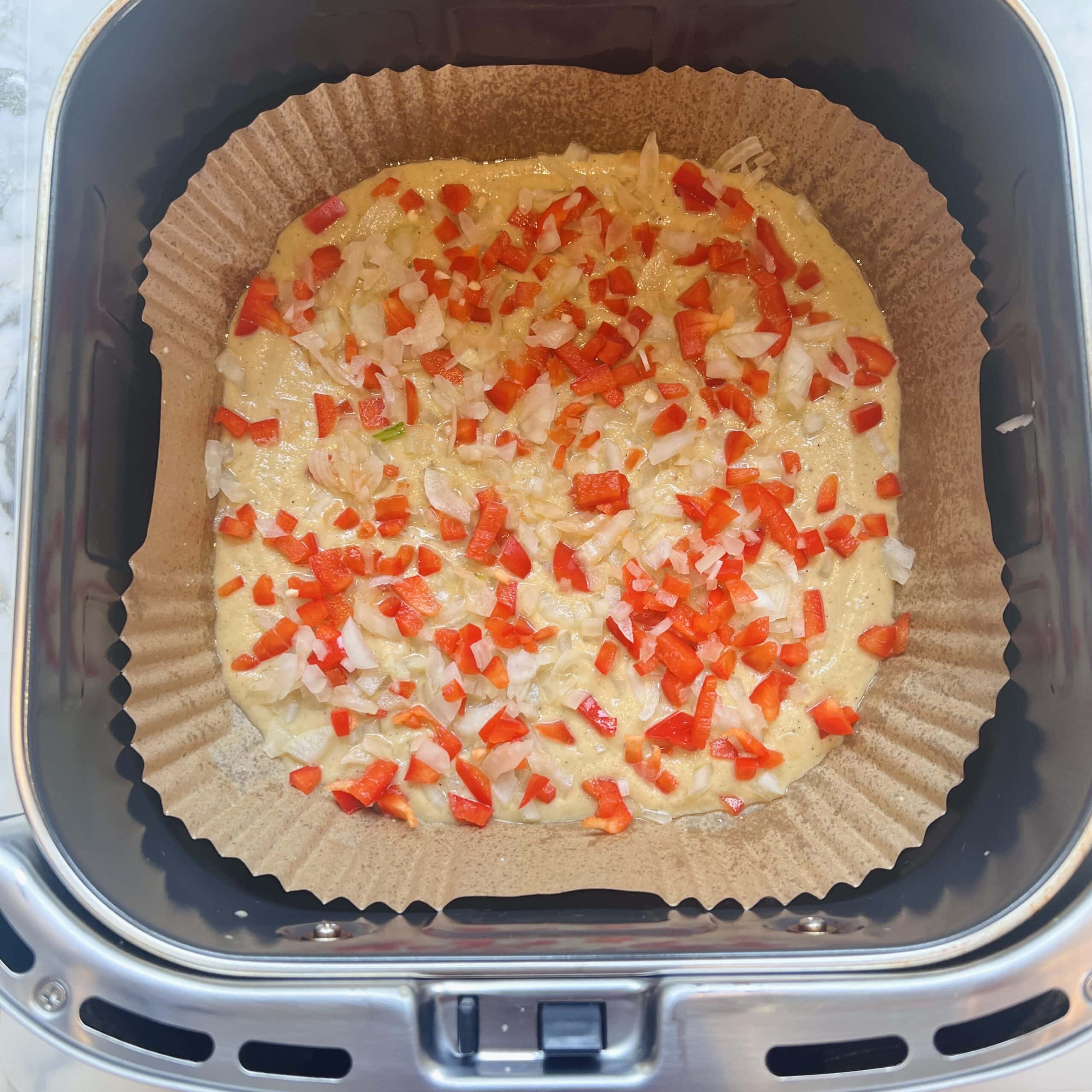 A baking dish lined with parchment holds uncooked batter topped with chopped red bell peppers and onions inside an air fryer.