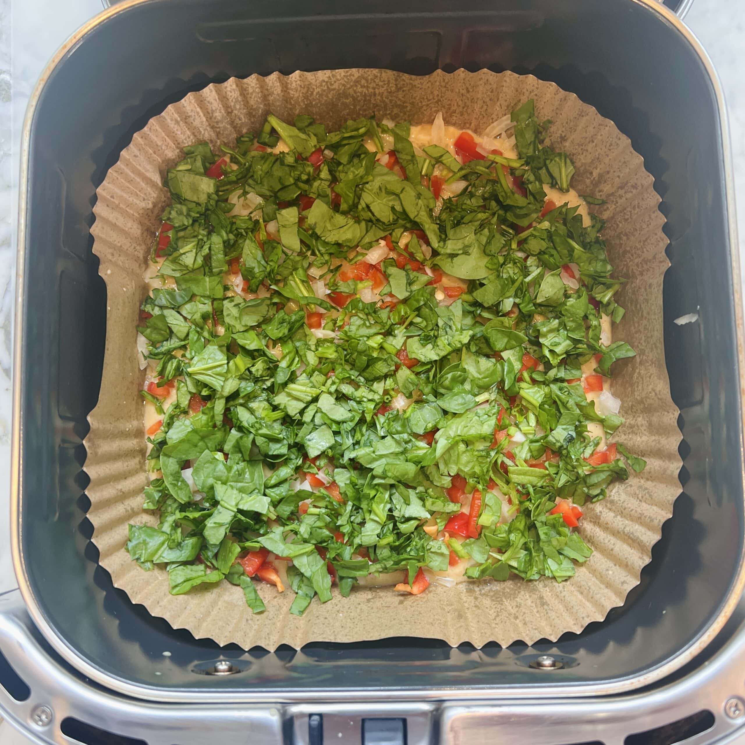 Chopped greens and diced red vegetables on parchment paper in an air fryer basket, ready to be cooked.