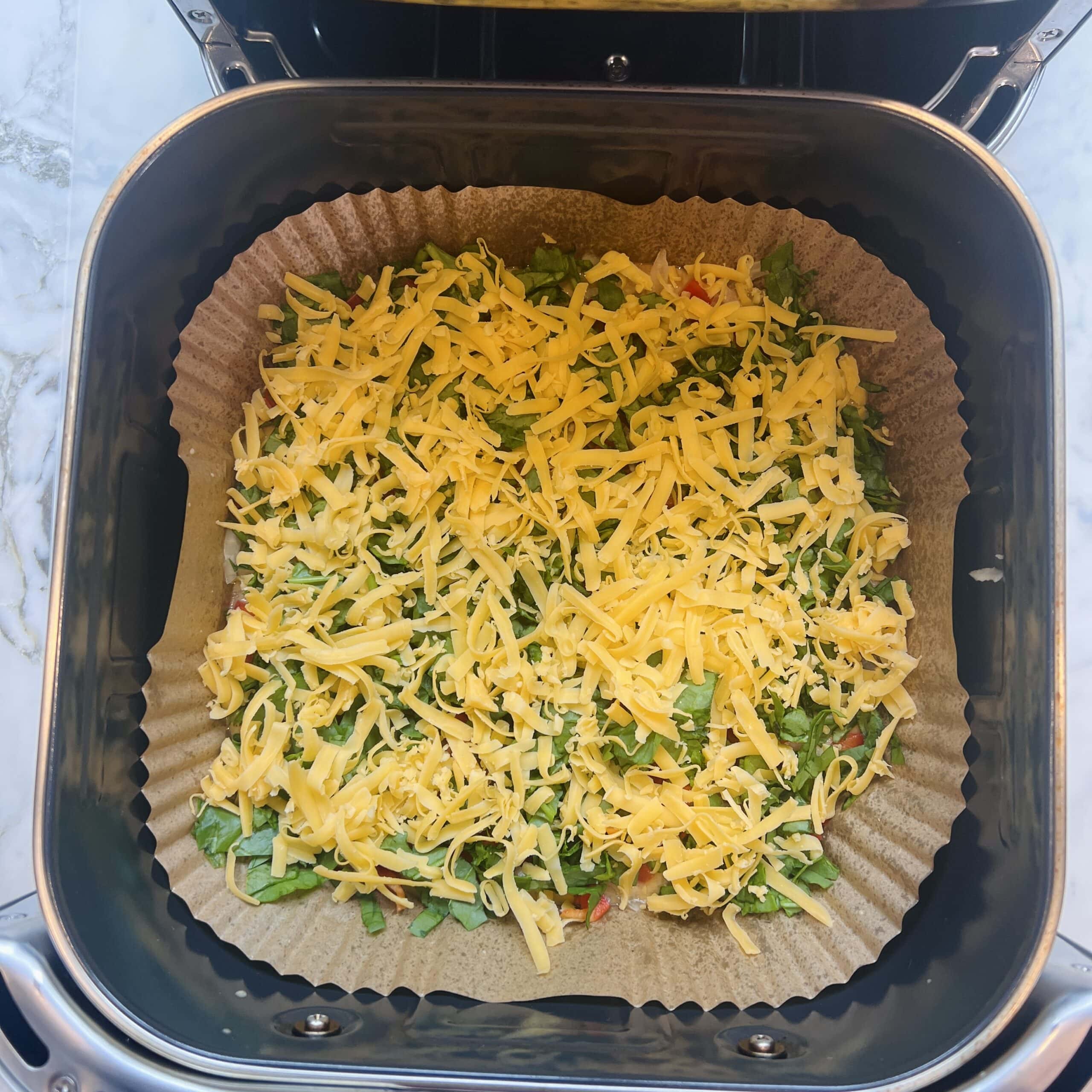 Grated cheese and chopped vegetables on parchment paper inside an air fryer basket, ready for cooking.