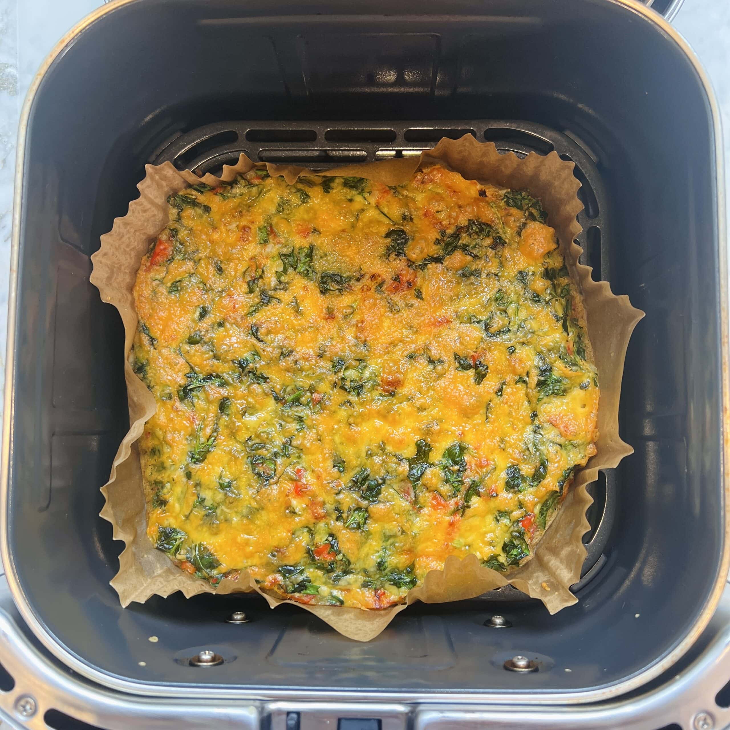 A cooked vegetable frittata sits on parchment paper inside an air fryer basket.