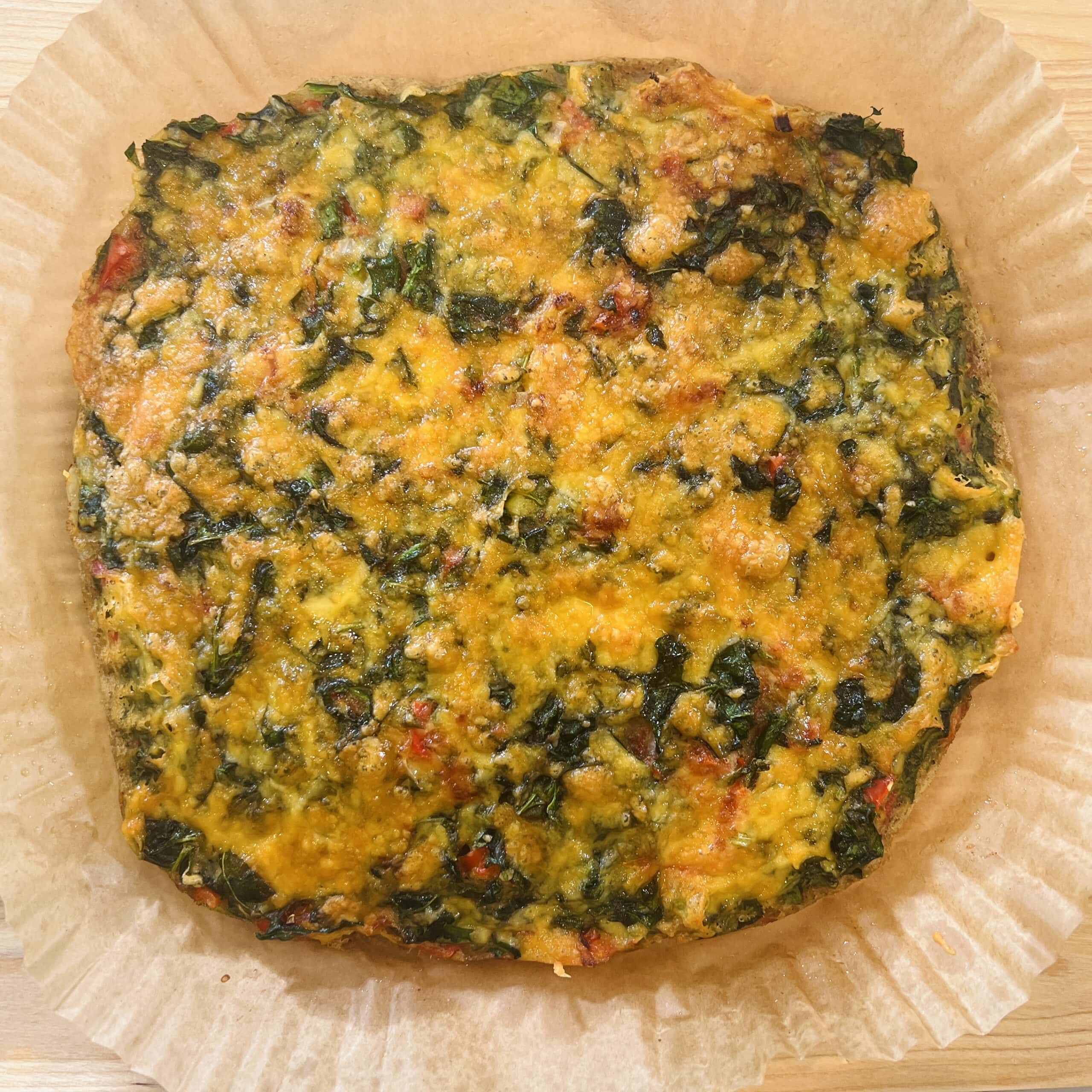 A baked vegetable frittata sits on parchment paper, showing a golden brown top speckled with greens and red peppers.