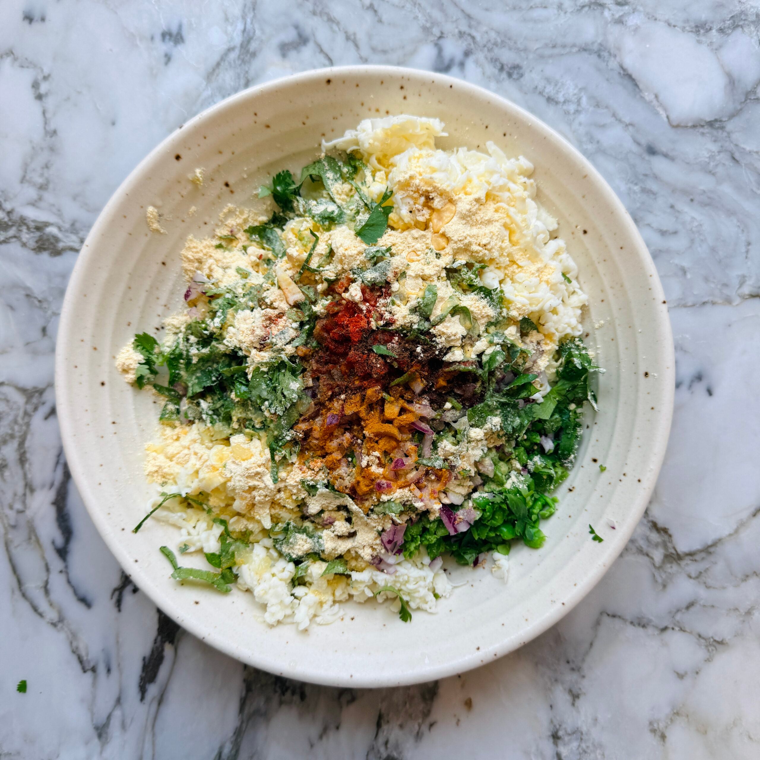 A bowl with crumbled paneer, chopped onions, cilantro, and assorted spices on a marble countertop.