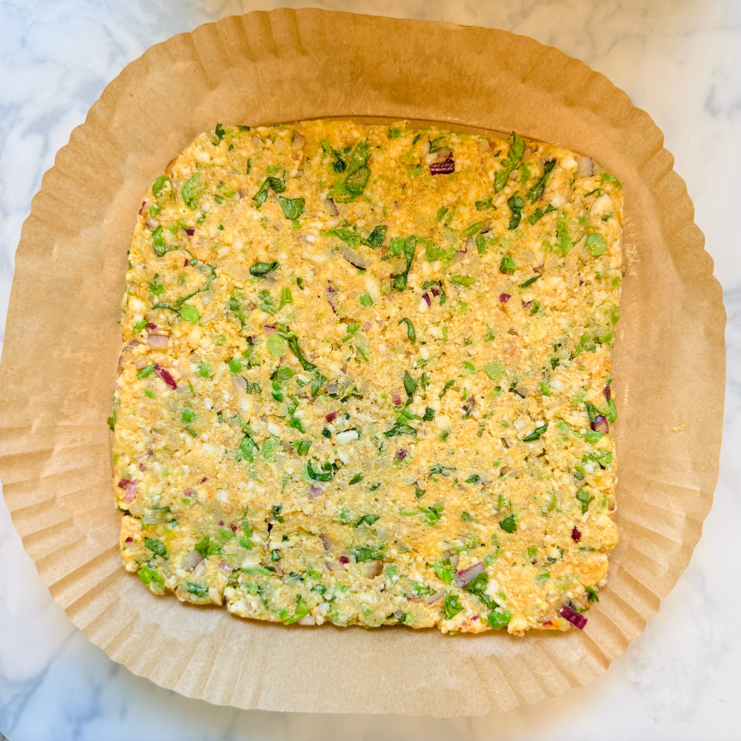A square, uncooked mixture of vegetables and batter is spread on parchment paper in a baking pan, ready to be baked.