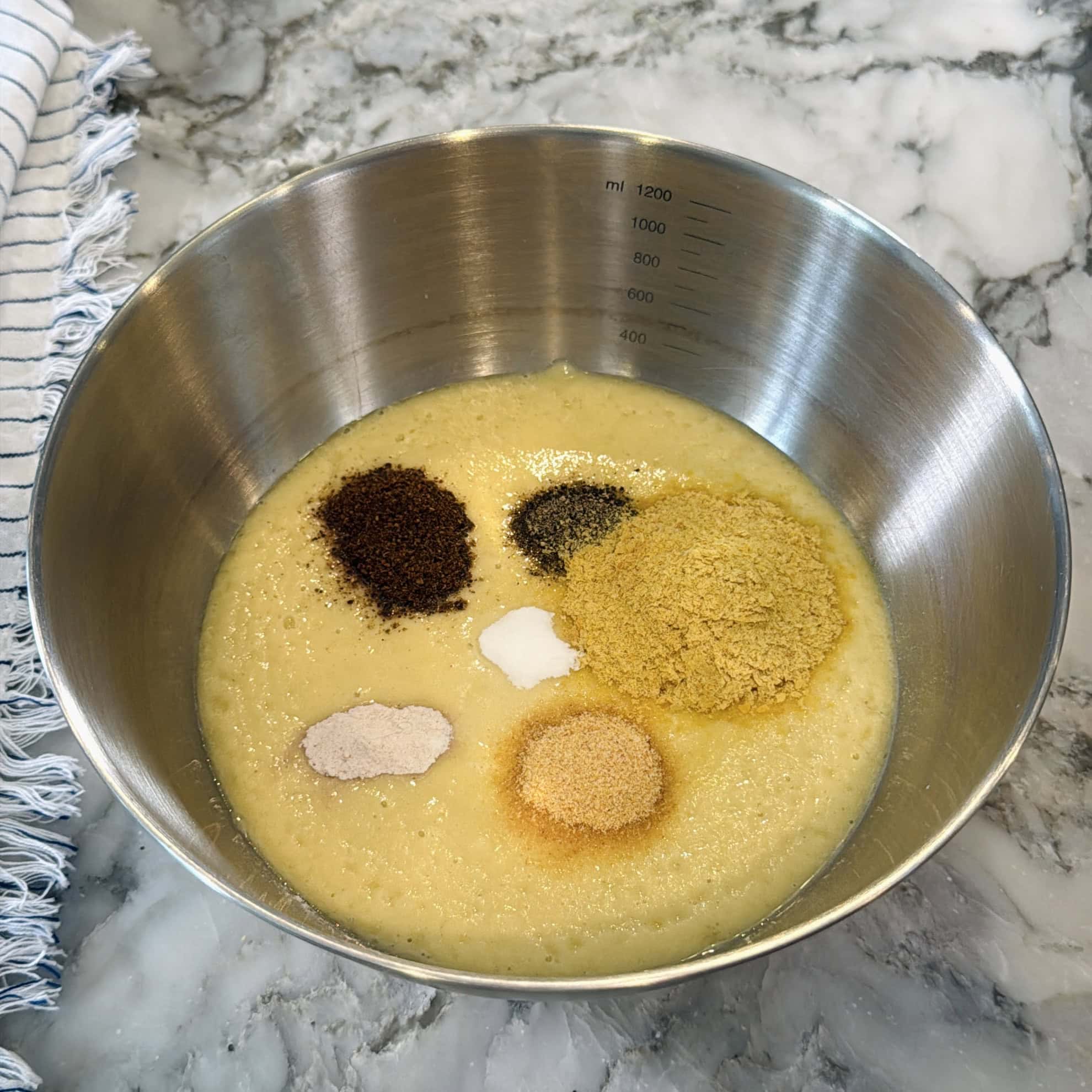 A stainless steel mixing bowl with a yellowish batter and six different seasonings on top, placed on a marble countertop next to a striped cloth.
