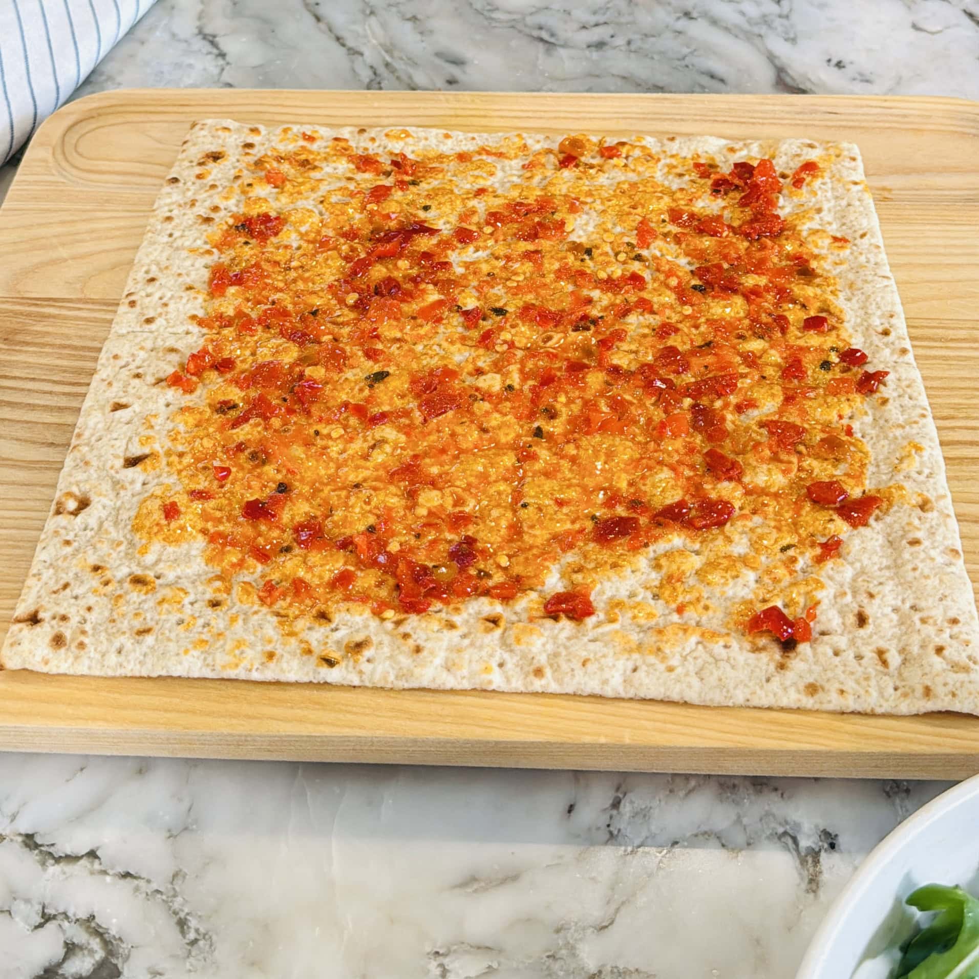 A square piece of flatbread spread with a layer of red pepper sauce sits on a wooden cutting board on a marble countertop.