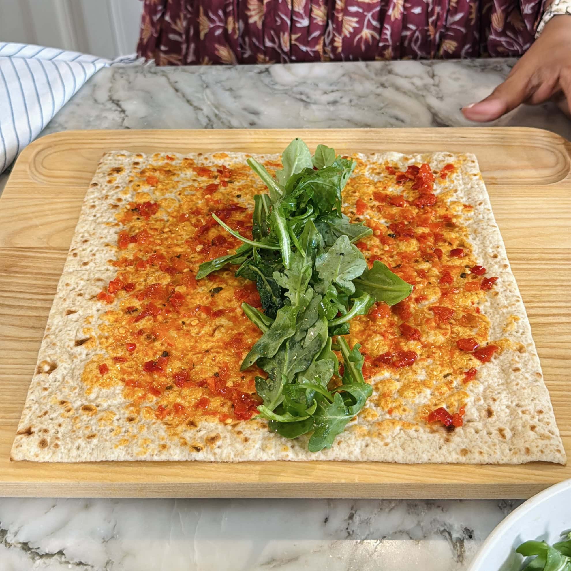 A square flatbread topped with red chili spread and a pile of fresh arugula, placed on a wooden board with a person seated behind it.