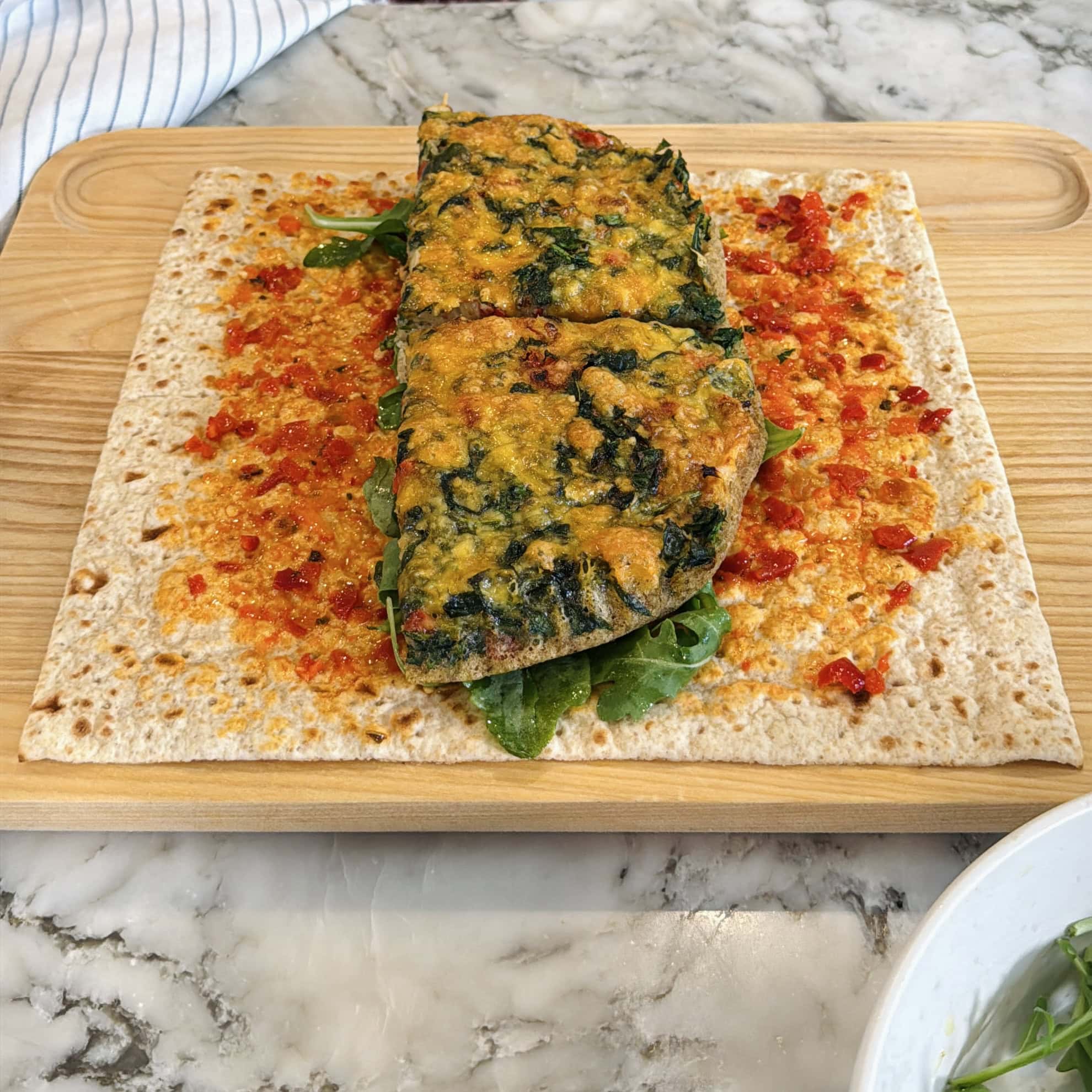 A folded spinach and cheese omelet on a flatbread with red chili sauce and greens, served on a wooden board.