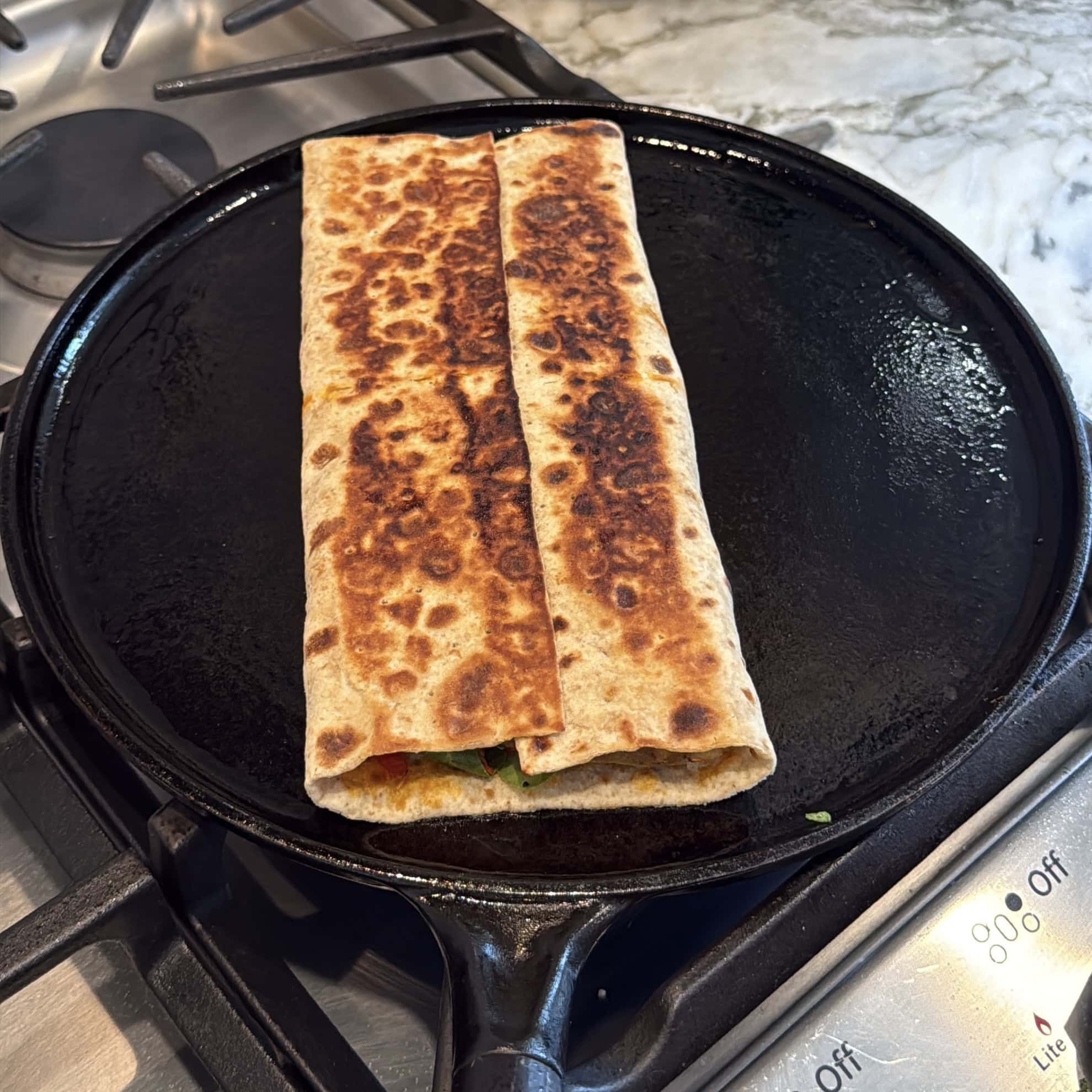 A folded, grilled flatbread wrap is being heated on a black cast iron pan over a stove.