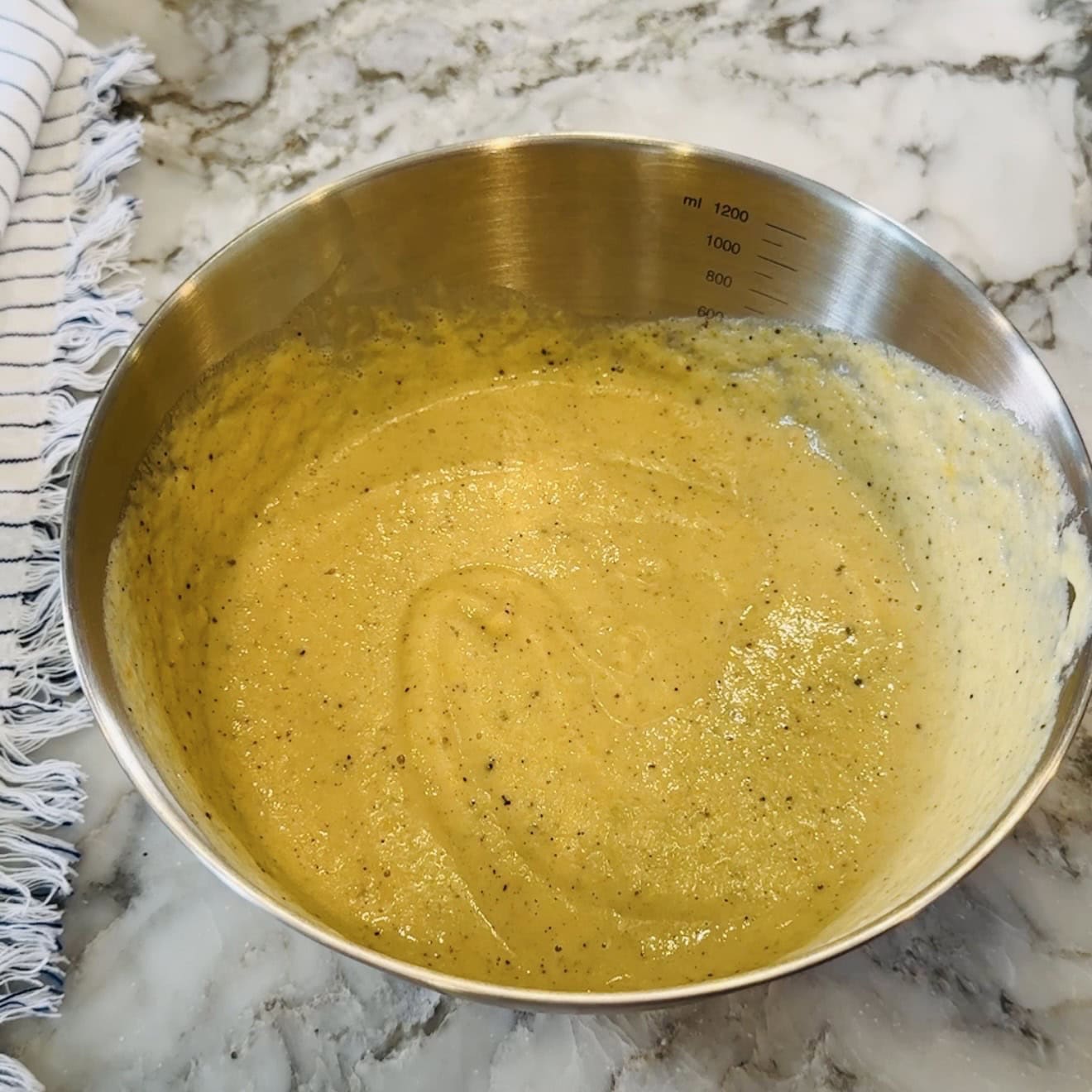 A stainless steel mixing bowl with yellow batter containing black specks sits on a marble countertop next to a striped cloth.