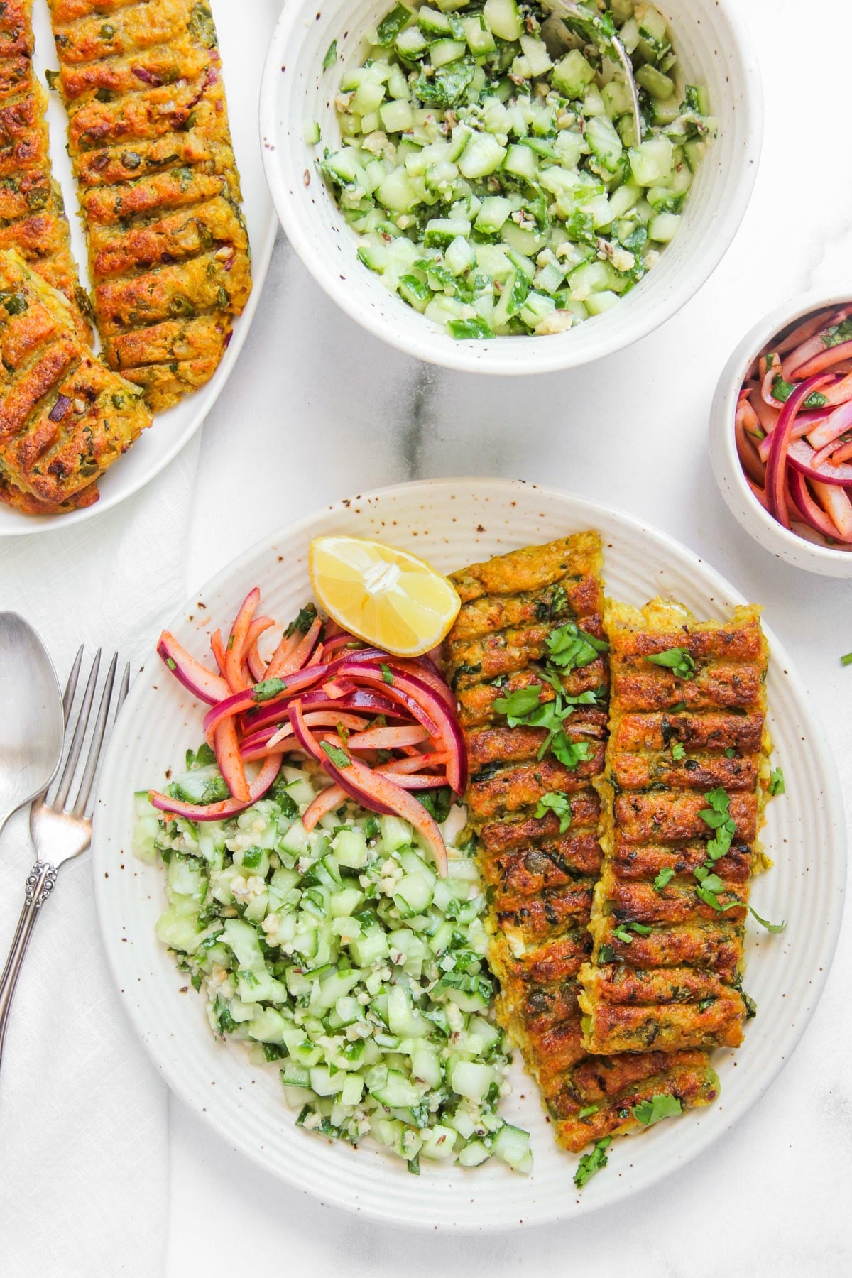 A plate with grilled kebabs, cucumber salad, and pickled onions, garnished with cilantro and a lemon wedge; additional salad bowls visible nearby.