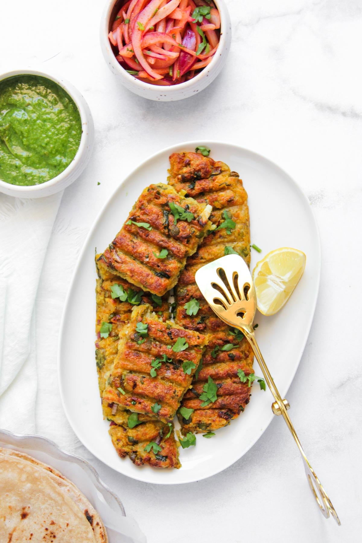 vegetarian sheet pan kebabs garnished with cilantro on a white plate, served with a lemon wedge, green chutney, pickled onions, and roti on the side.