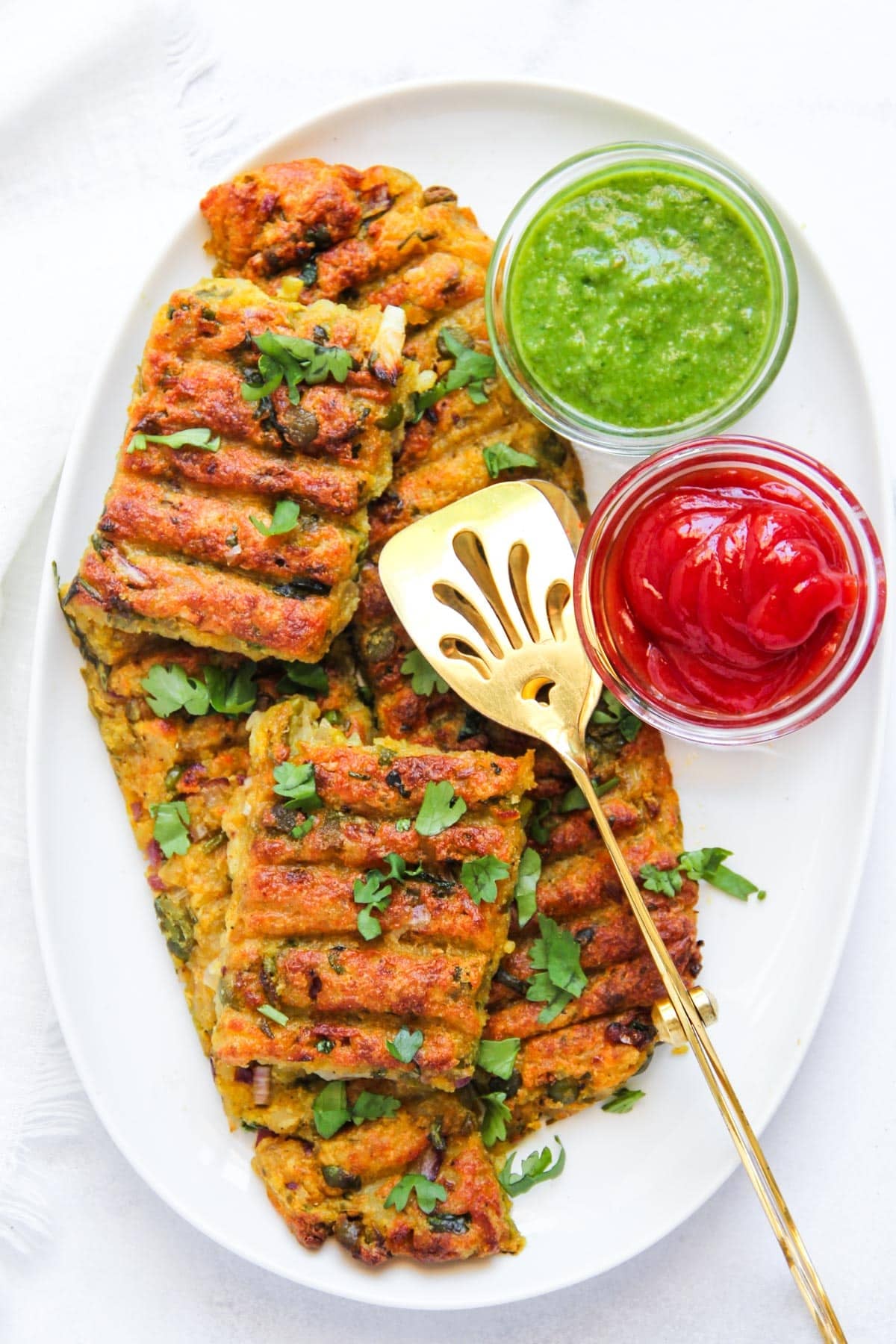 vegetarian seekh kebas garnished with cilantro, served on a white plate with green chutney and ketchup in small glass bowls, alongside a gold slotted serving spatula.