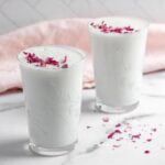 Two glasses of sweet lassi topped with dried rose petals, placed on a marble surface with a pink cloth in the background.