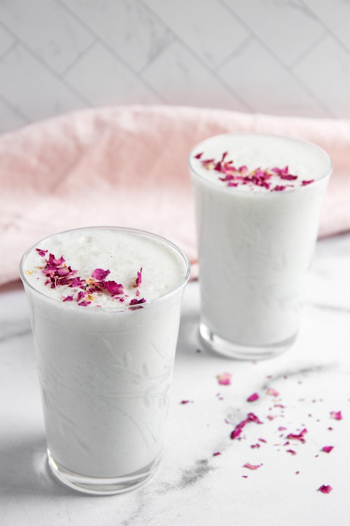 Two glasses of white, frothy lassi are topped with dried rose petals.