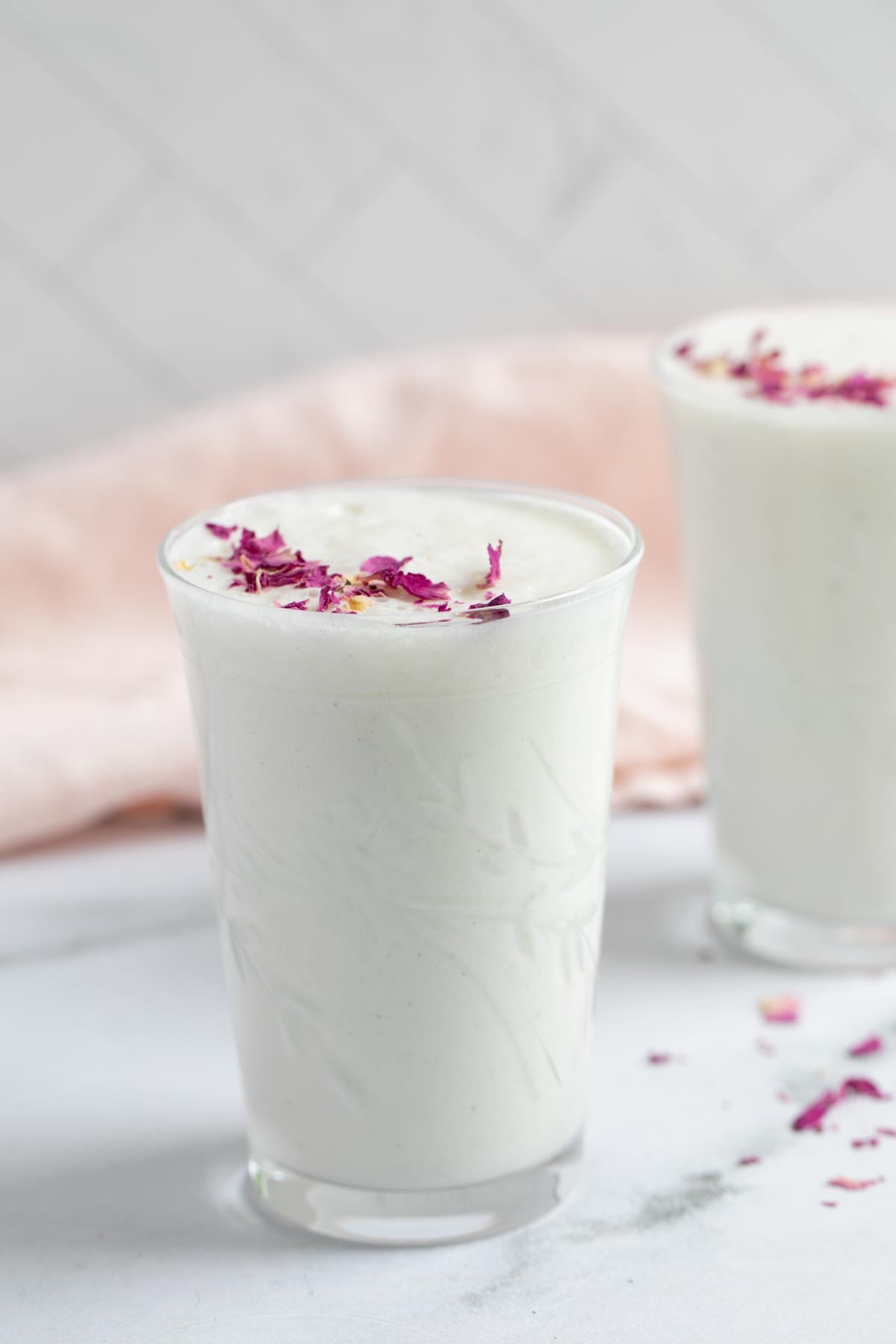 Two glasses of frothy-white sweet lassi topped with dried rose petals.