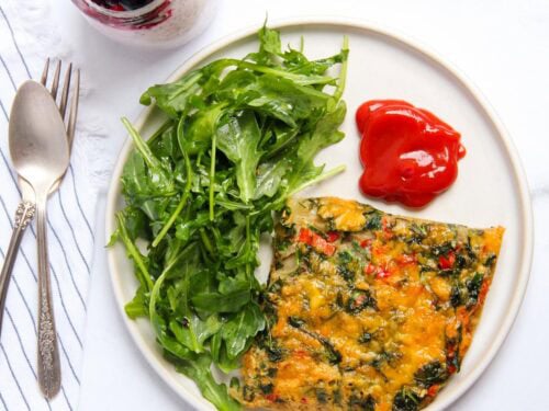 A plate with a slice of vegetable frittata, a serving of arugula, and two dollops of ketchup. A spoon, fork, and a glass of berry yogurt parfait are beside the plate.