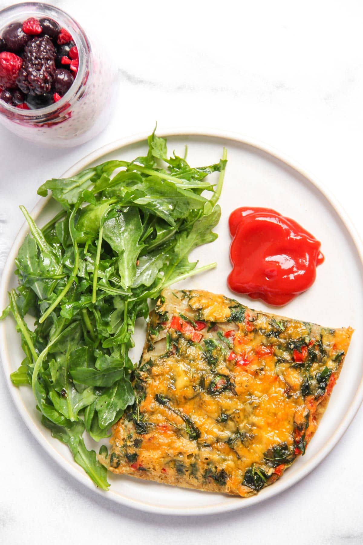 A plate with a slice of vegetable frittata, a serving of arugula salad, and a portion of ketchup; a jar of mixed berries and yogurt is placed next to the plate.