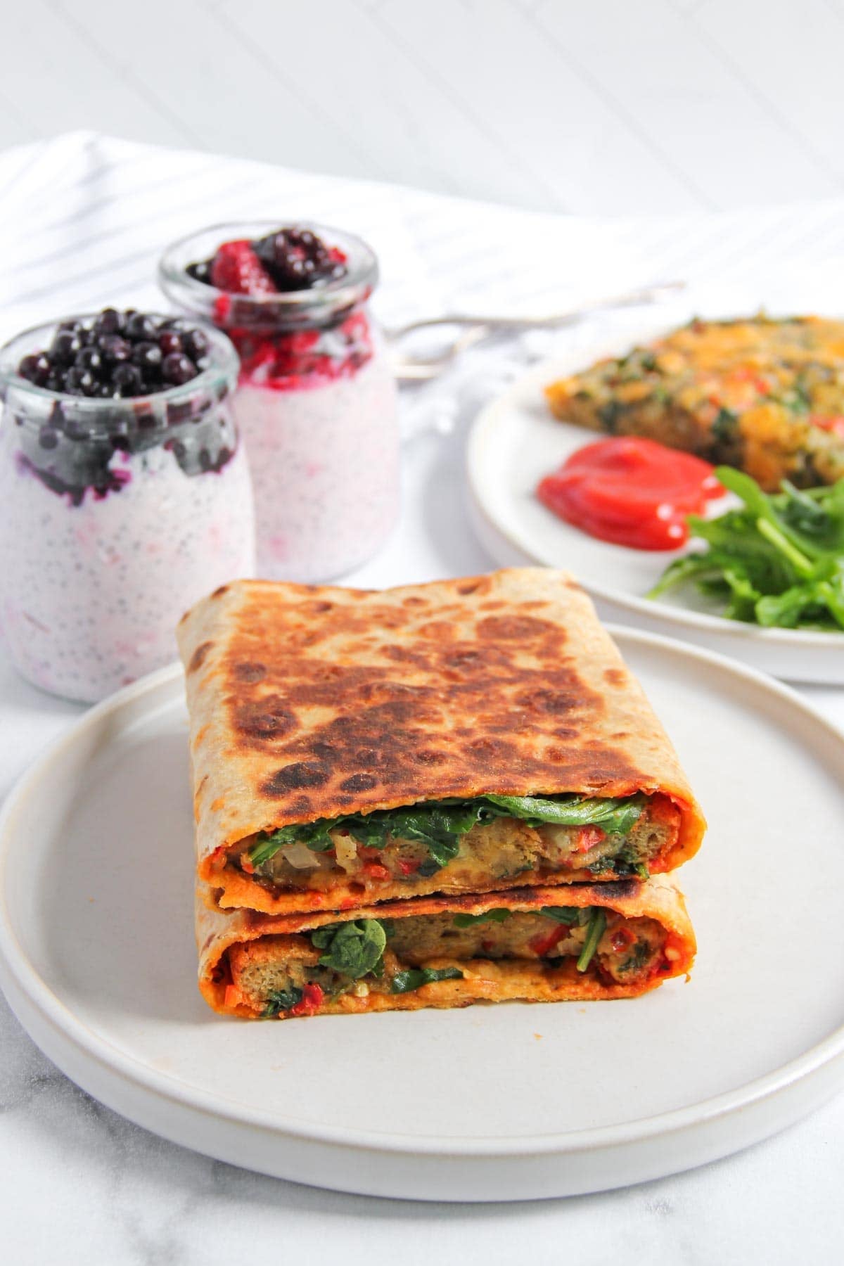 A plate with a grilled wrap cut in half, filled with vegetables. In the background are two jars of yogurt with berries and a plate with greens and a slice of casserole.
