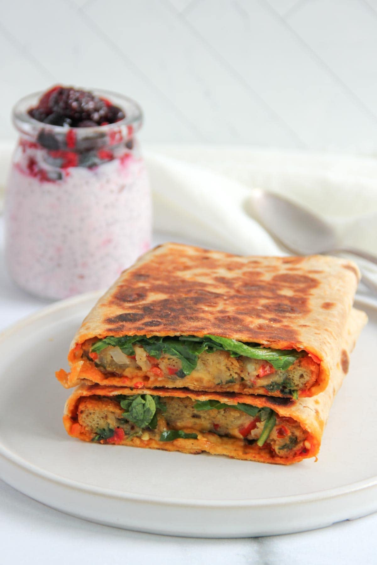 A folded, grilled wrap filled with vegetables, sliced in half and stacked on a plate, with a jar of yogurt topped with berries in the background.