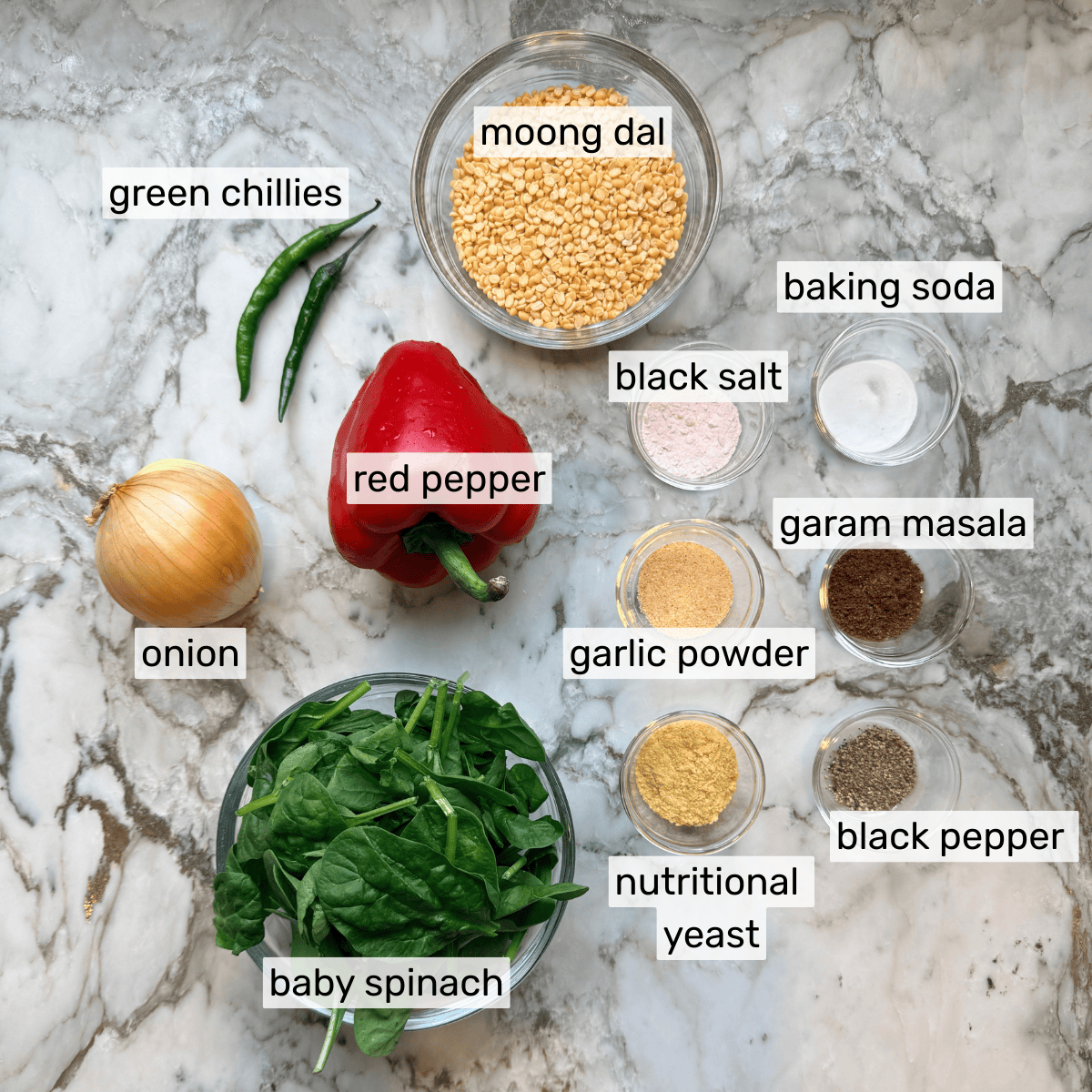 A marble surface with labeled ingredients: green chillies, moong dal, baking soda, black salt, garam masala, garlic powder, black pepper, nutritional yeast, baby spinach, onion, and red pepper.