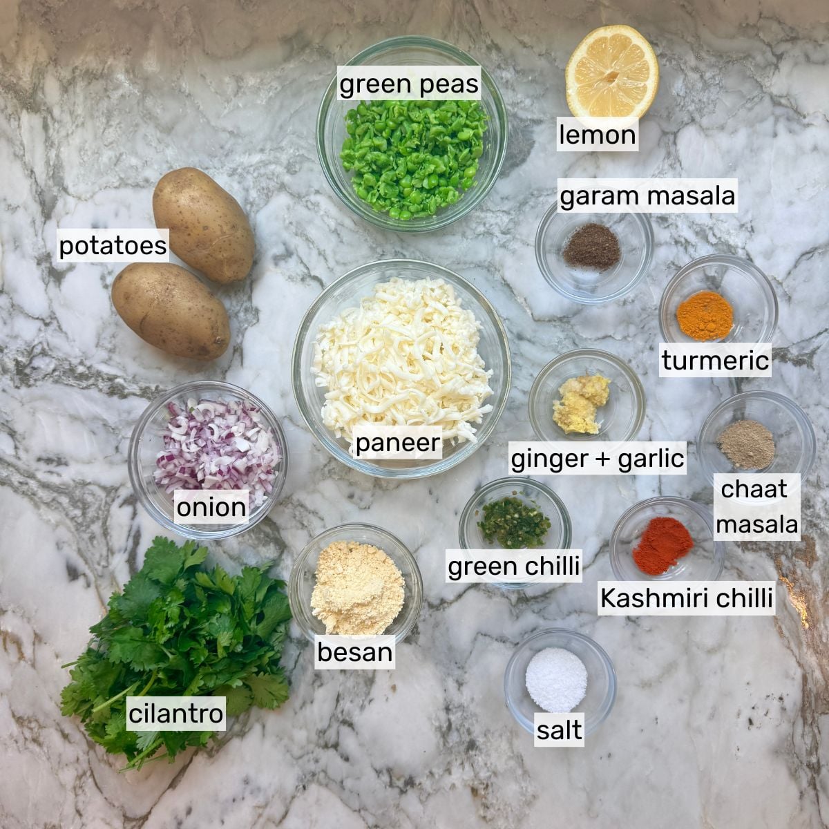 Ingredients for vegetarian sheet pan kebabs arranged on a marble surface, including potatoes, paneer, green peas, lemon, onion, cilantro, spices, besan, salt, and ginger-garlic paste in labeled bowls.