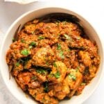 A bowl filled with Chicken Sukka, an Indian chicken dish garnished with chopped herbs, placed on a light-colored surface.