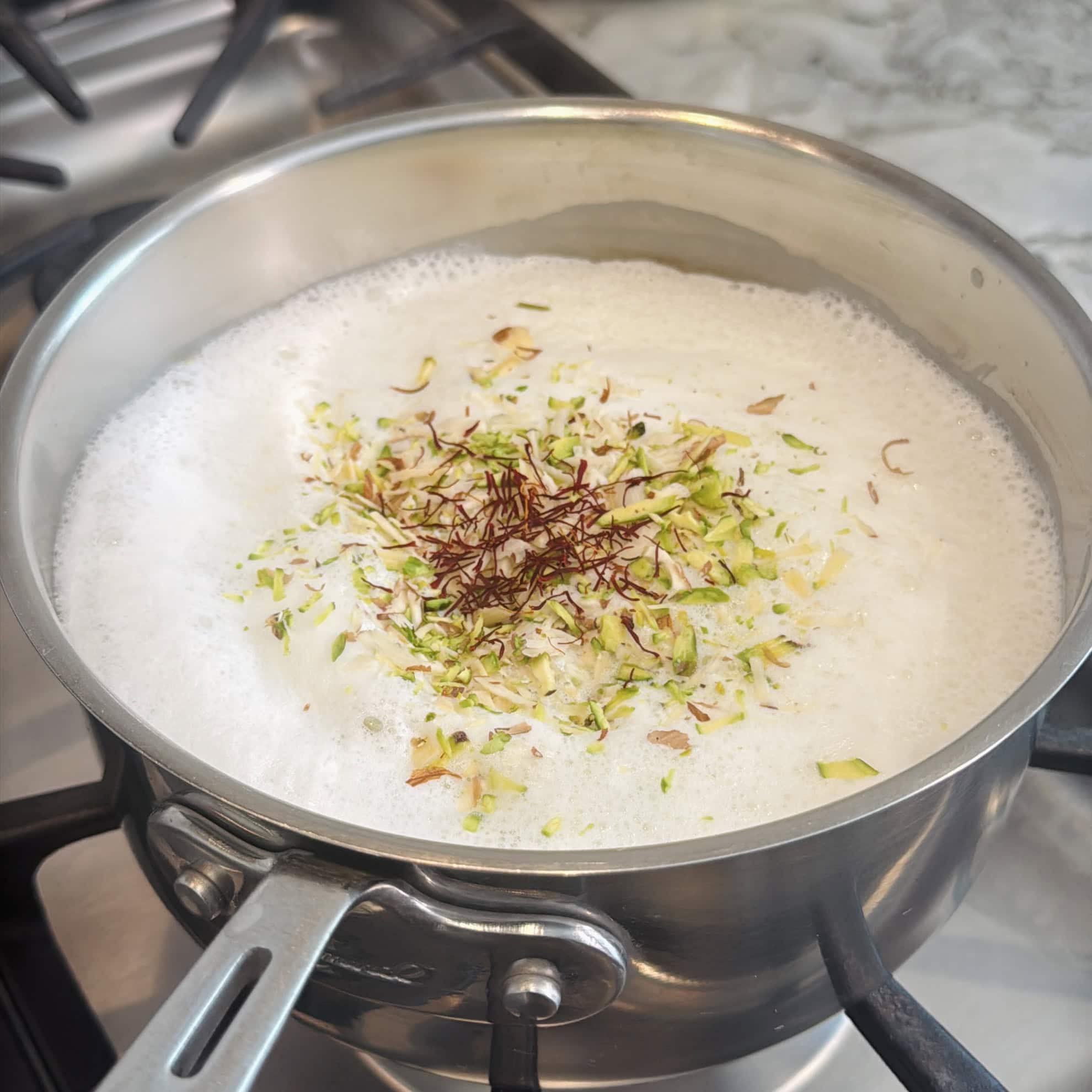 A saucepan of boiling milk on a stove, topped with saffron strands, chopped pistachios, and almonds.