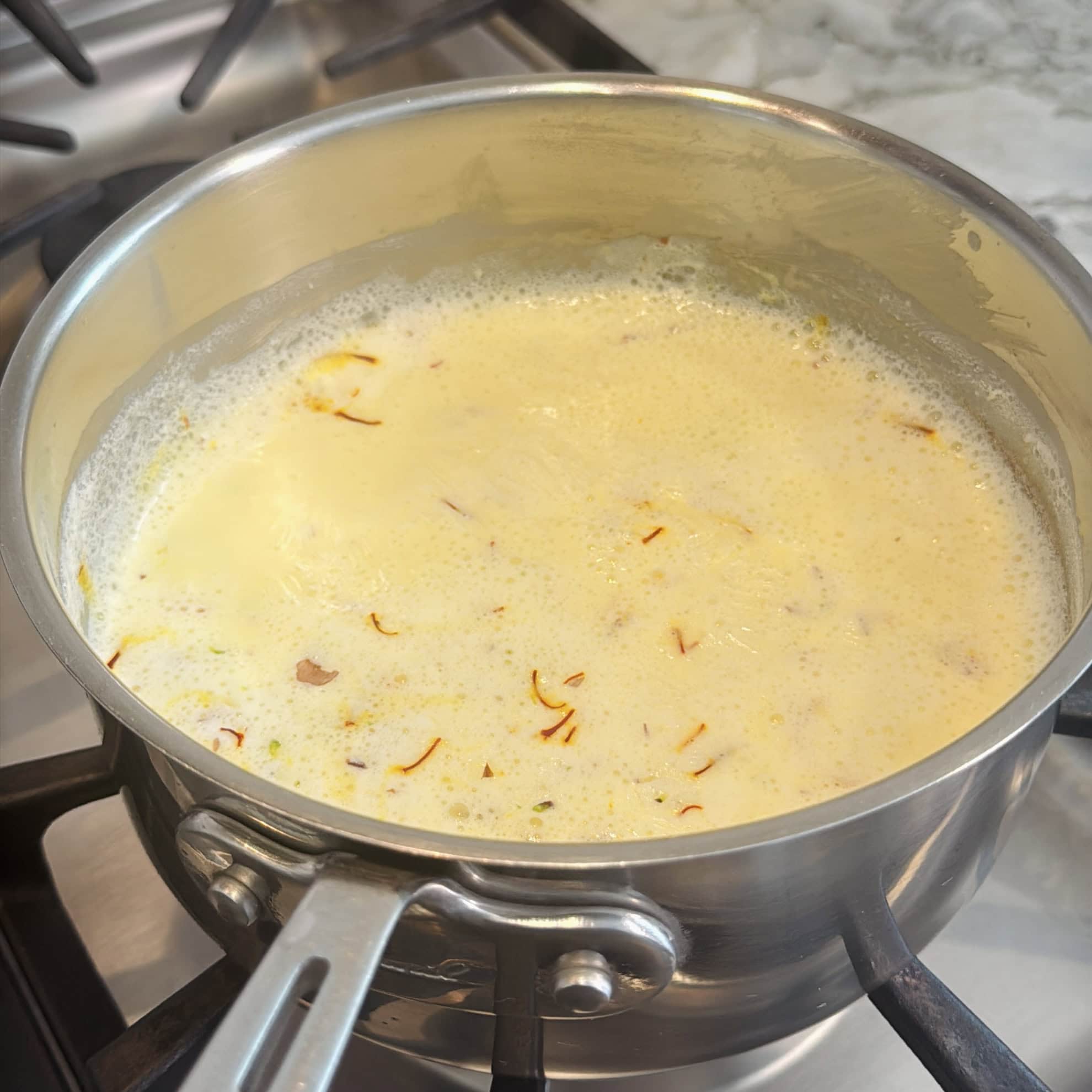 A stainless steel pot on a stove contains boiling milk with saffron strands and some spices visible on the surface.