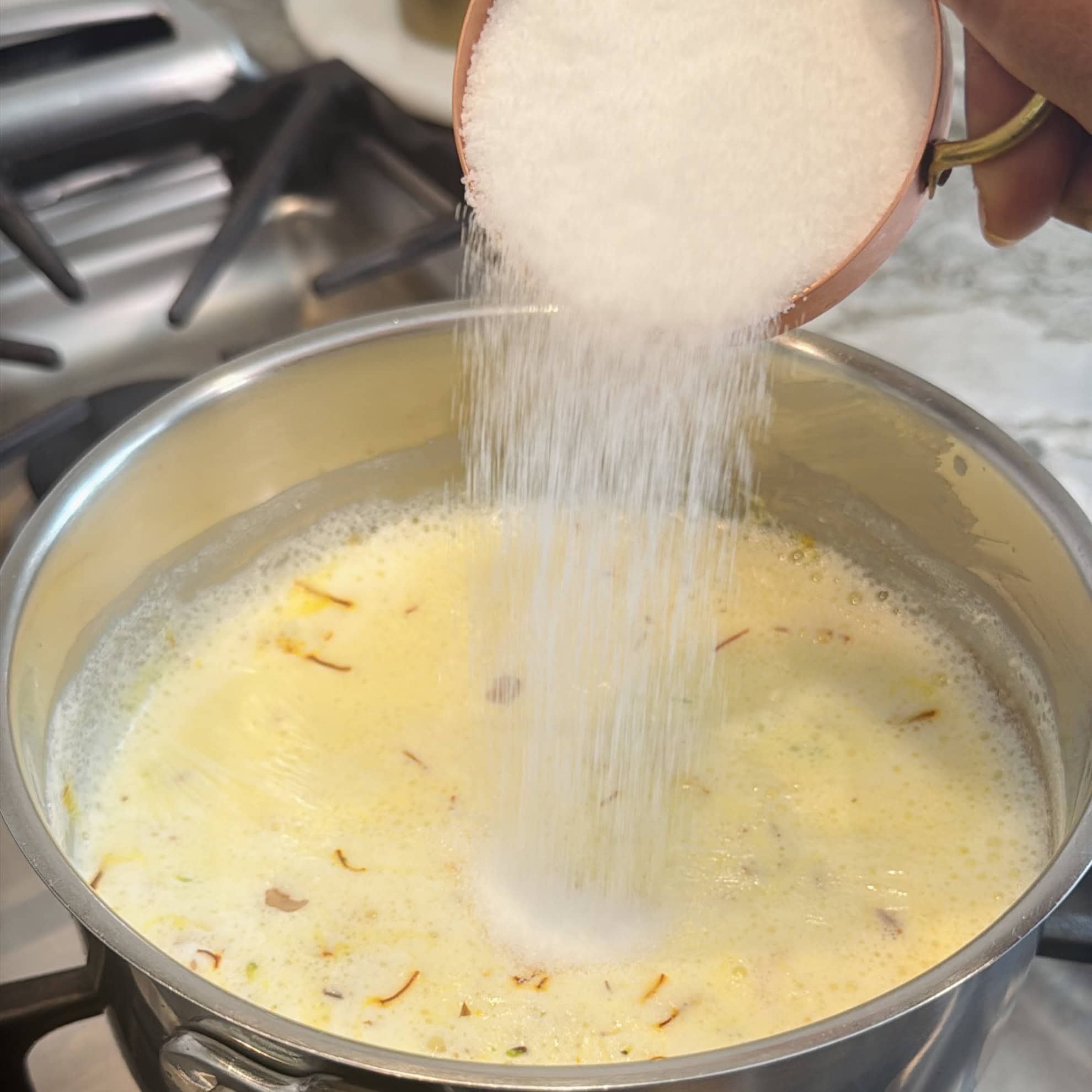 A hand pours a cup of white sugar into a pot of simmering yellow milk with saffron strands on a stovetop.