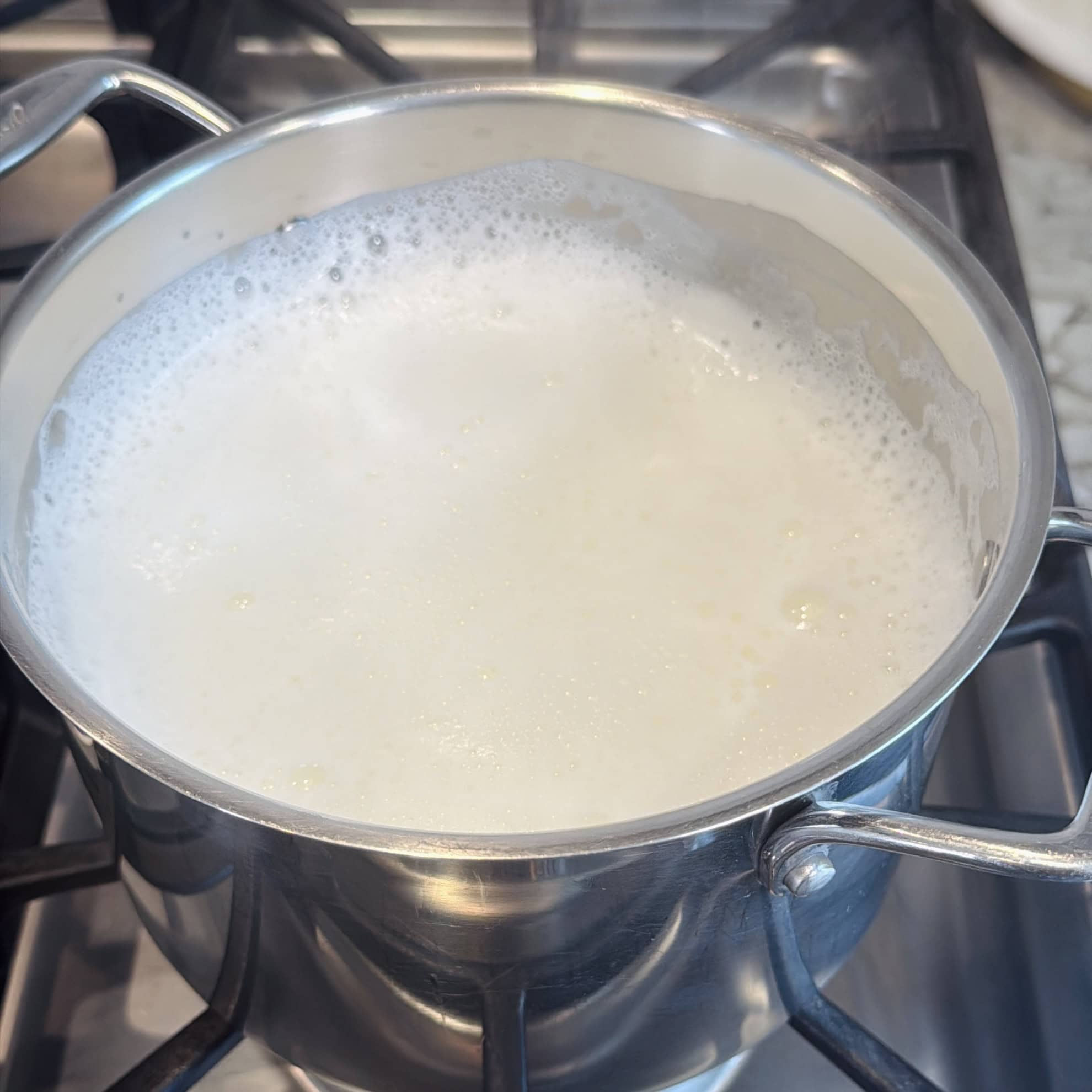 A stainless steel pot filled with milk is heating on a gas stove, with foam and bubbles forming on the surface.