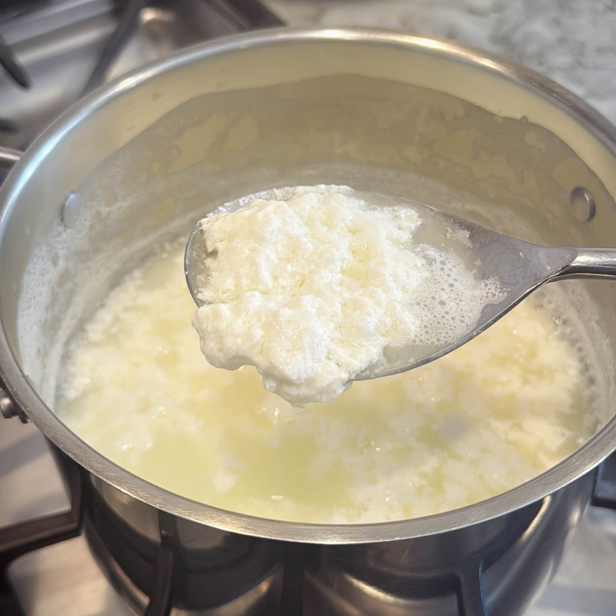 A metal spoon holds homemade ricotta cheese curds over a pot of whey on a stovetop.