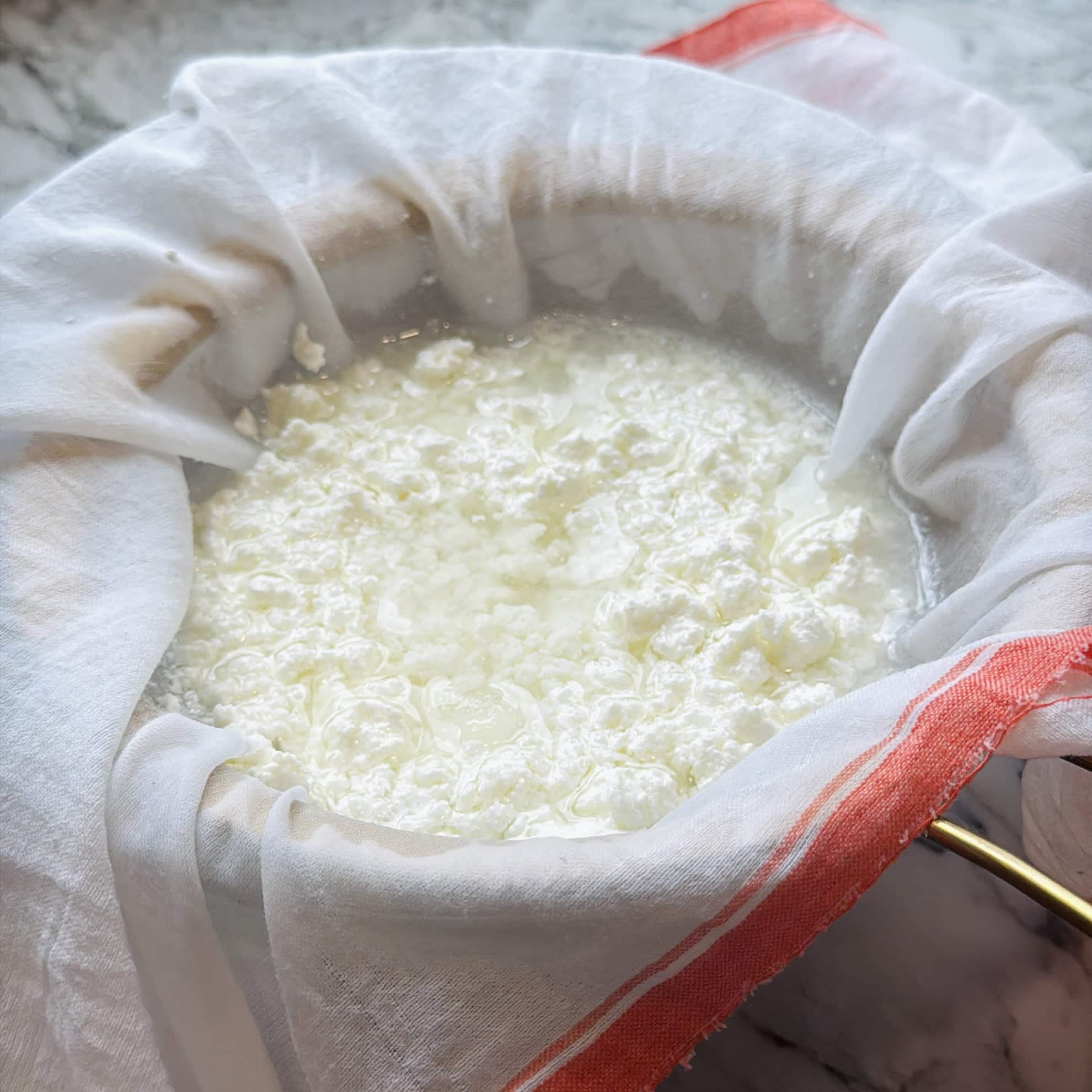 Curds and whey being strained through a cheesecloth-lined sieve, placed over a bowl on a marble countertop.