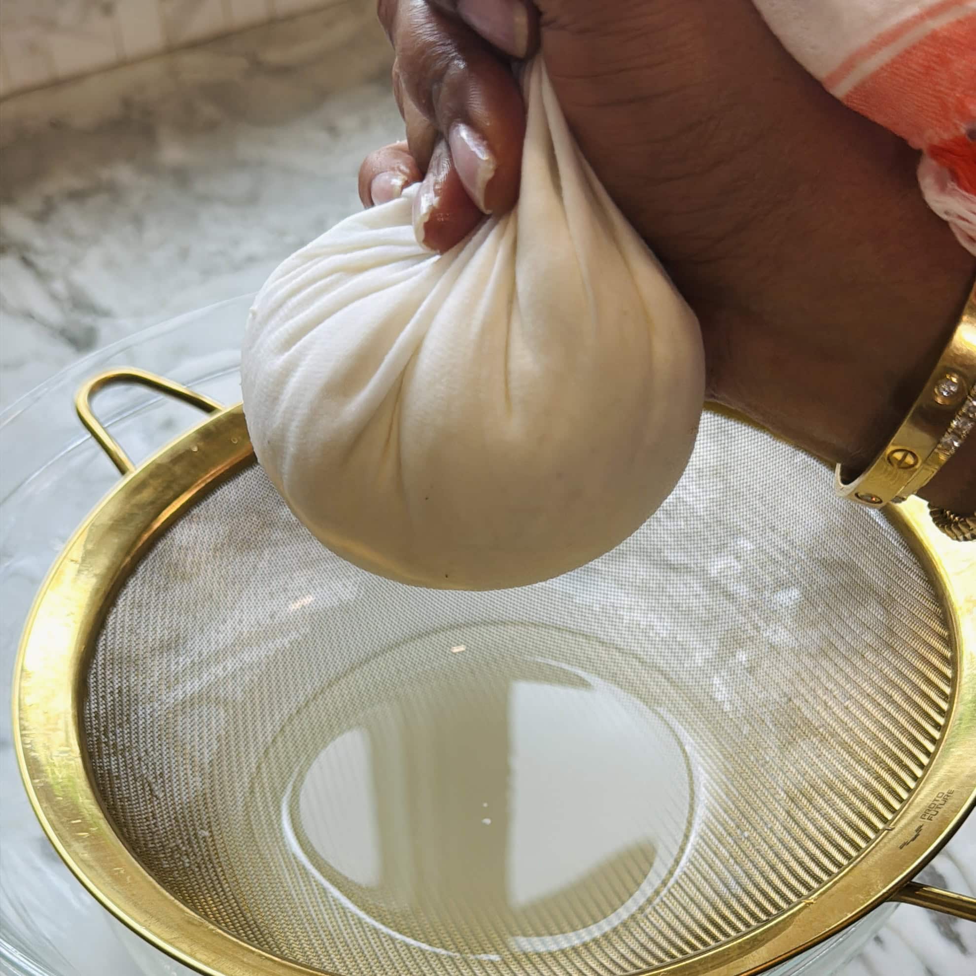 A hand squeezes a cloth bundle of cheese over a strainer, extracting liquid into a glass bowl below on a marble countertop.