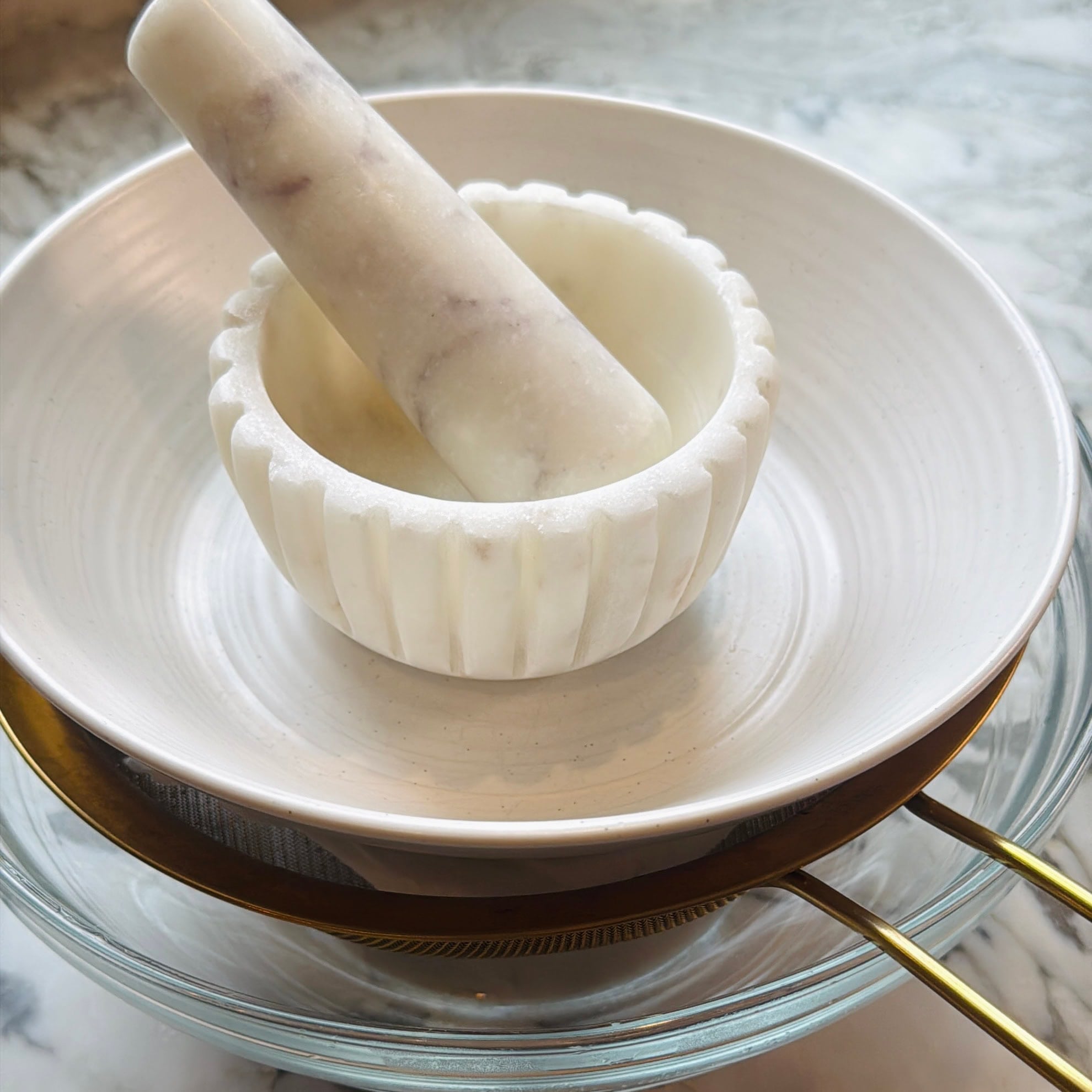A white marble mortar and pestle placed in a white bowl, which is stacked on top of a glass bowl and a gold-colored fine mesh strainer.